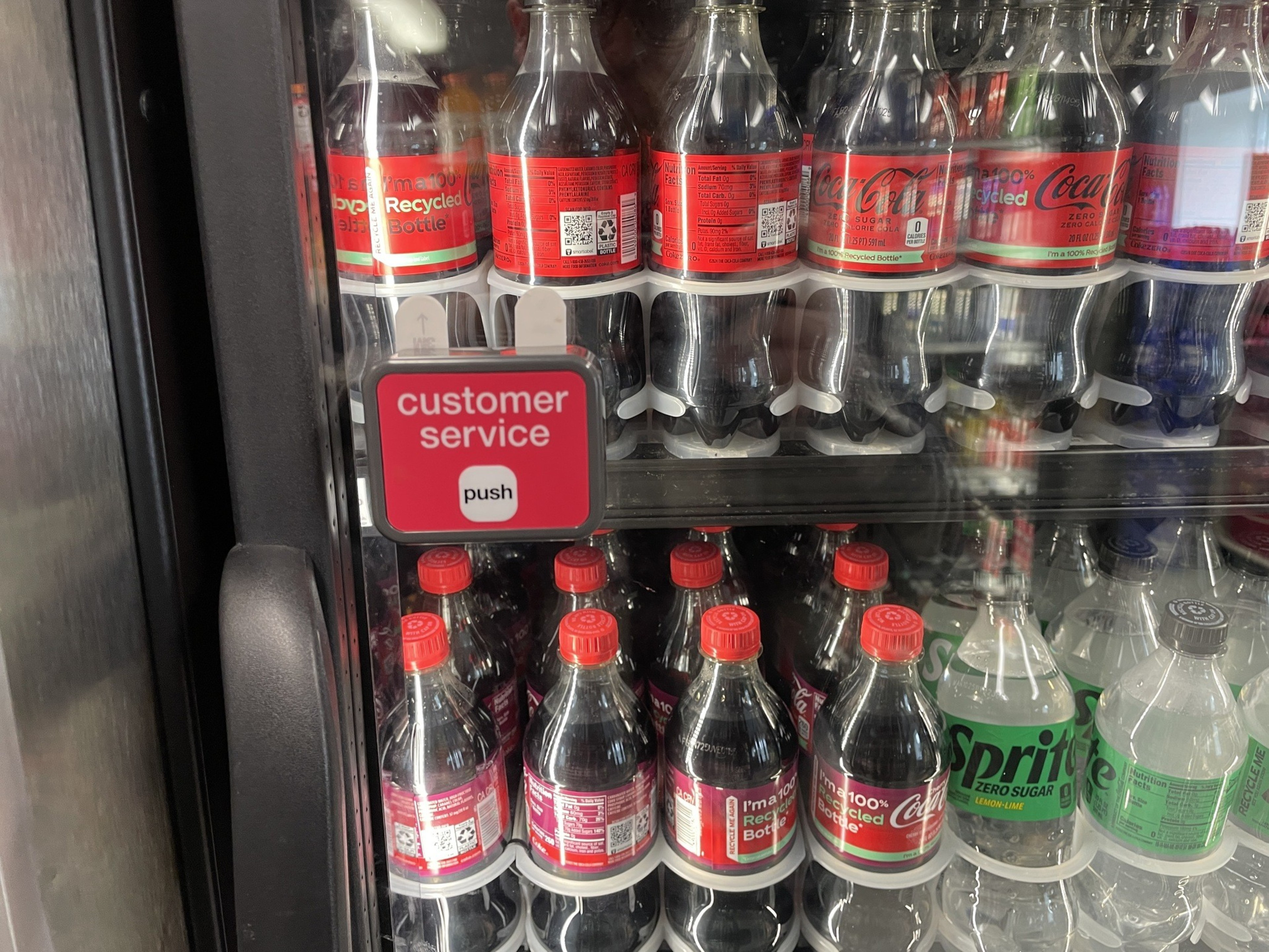 A cooler showcases rows of Coca-Cola and Sprite bottles. A &quot;customer service&quot; button labeled &quot;push&quot; is attached to the cooler door.