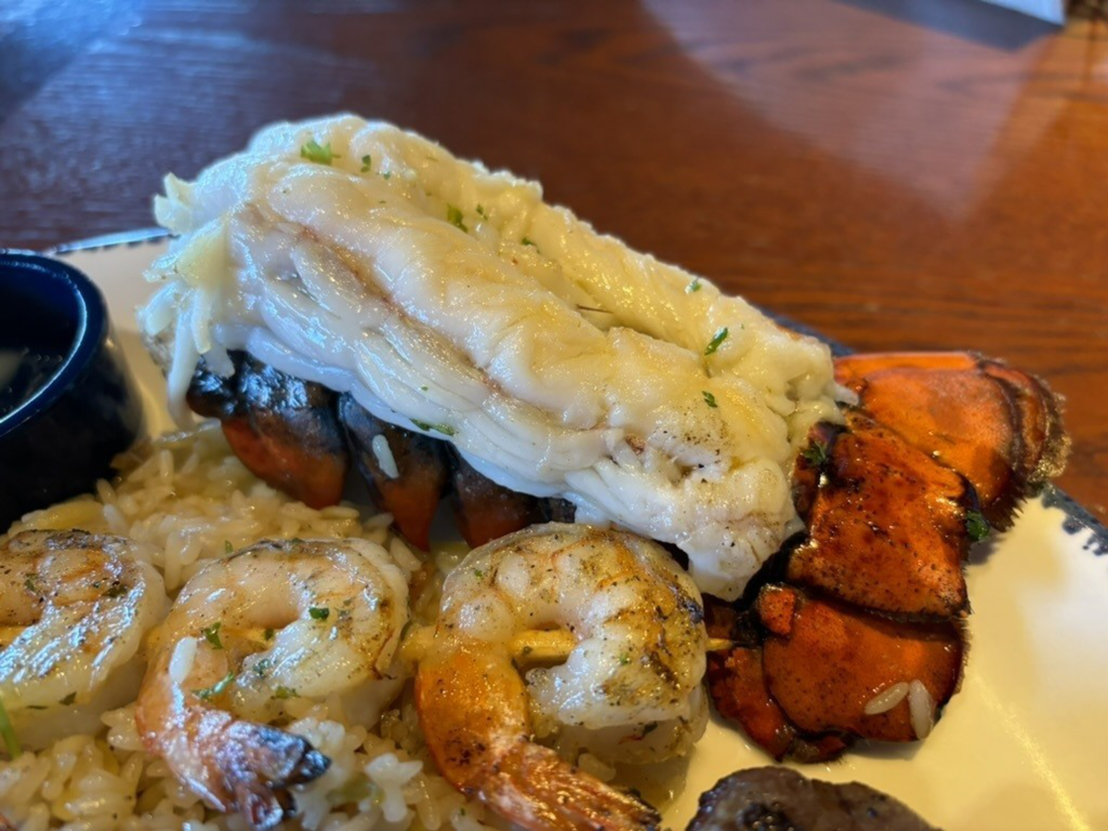 The image shows a close-up of a seafood dish featuring a lobster tail and grilled shrimp on a bed of rice, garnished with herbs, accompanied by a small dish of sauce.