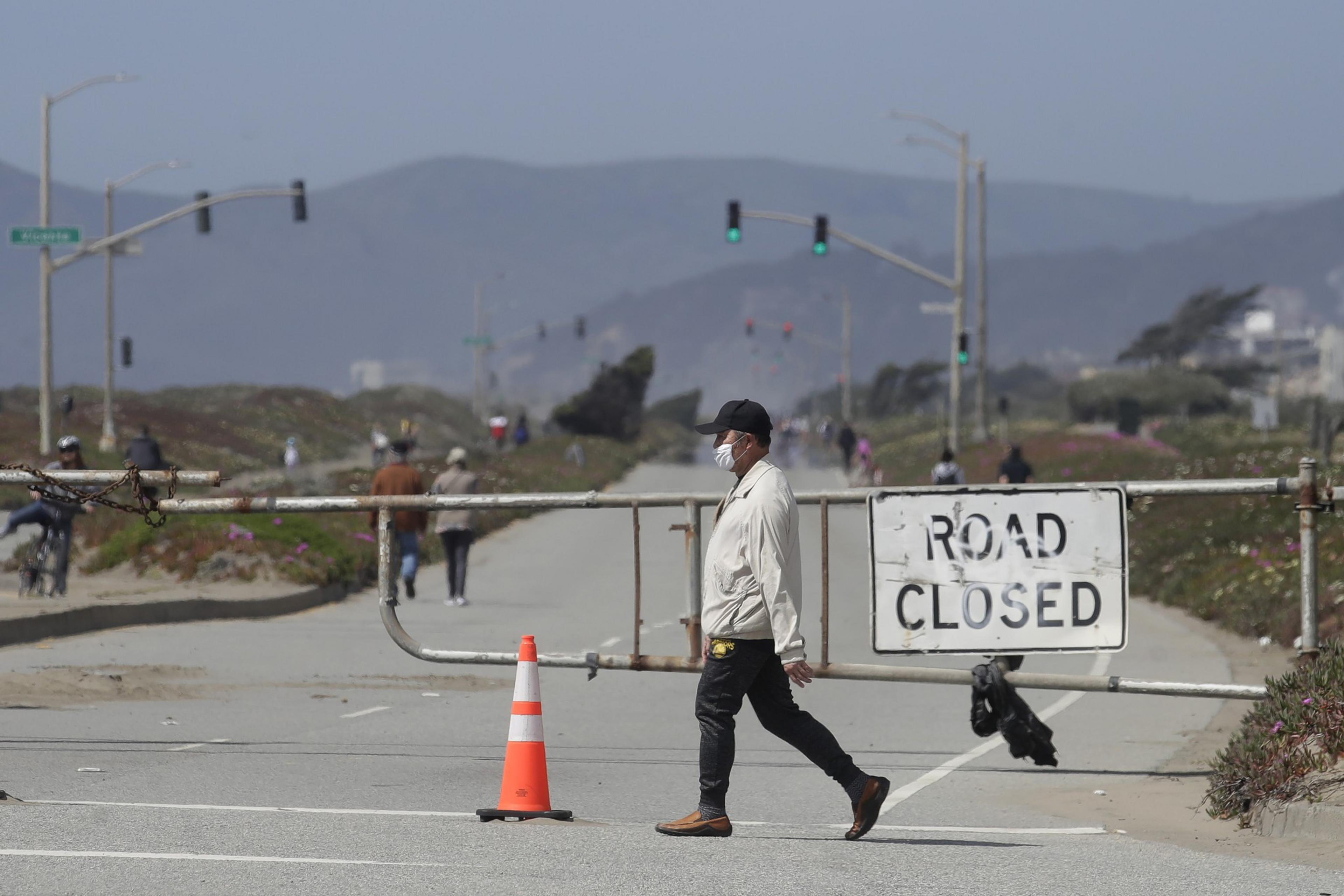 A man wearing a mask walks across a street closed off by a gate and a &quot;Road Closed&quot; sign. In the background, people are walking, and mountains are visible.