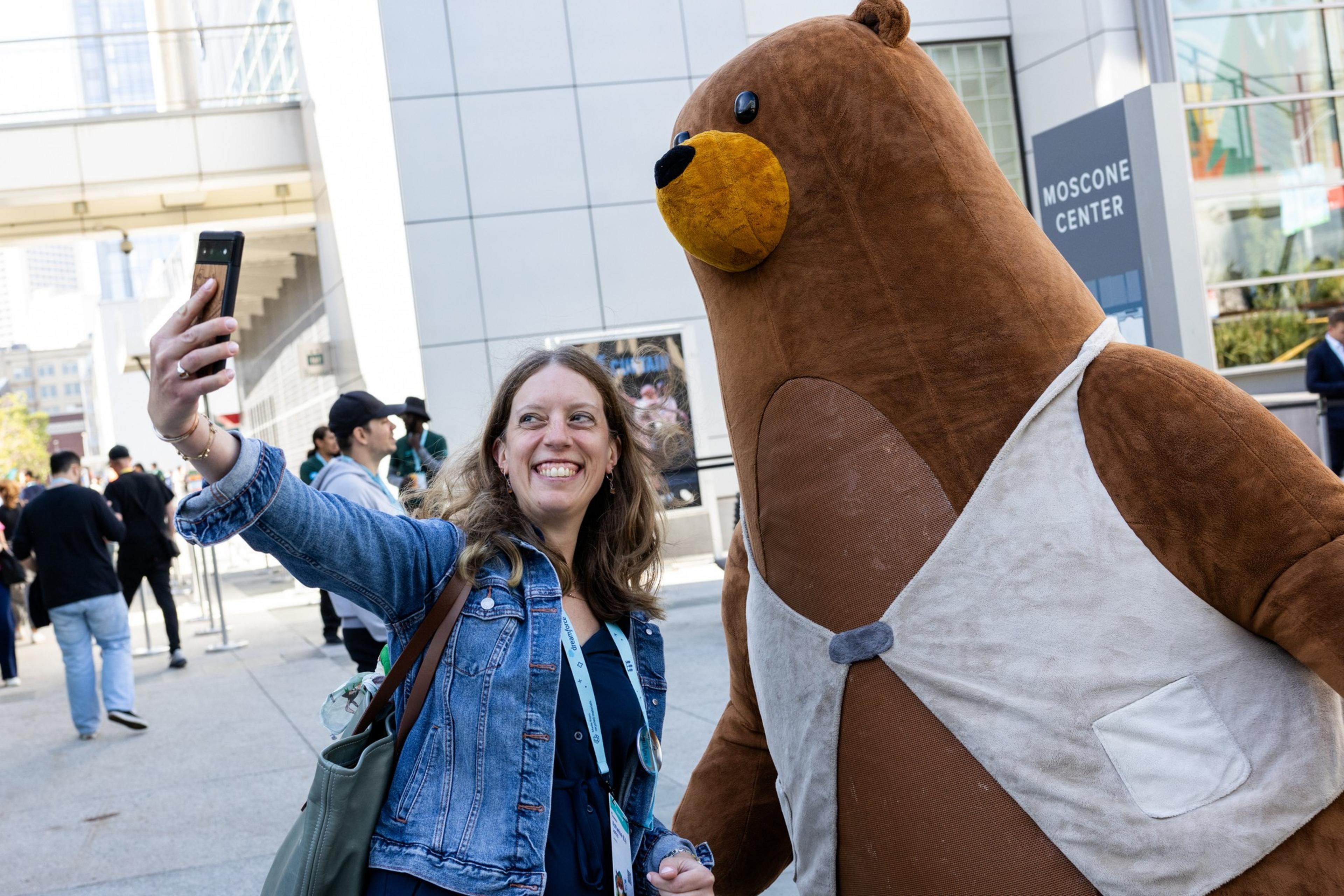 A woman smiles while taking a selfie with a person in a large brown bear costume outside the Moscone Center.