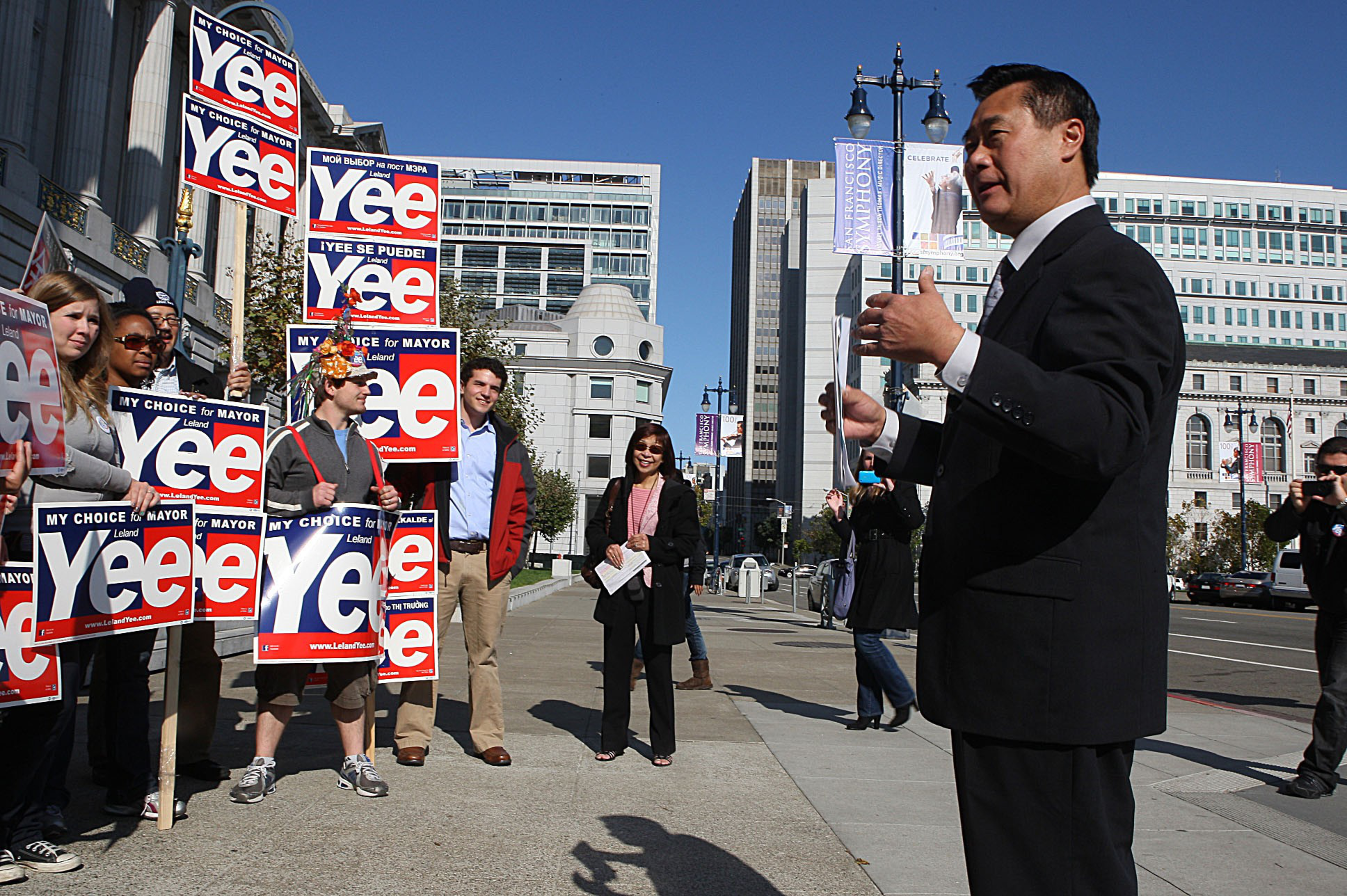 A group of people holding "Yee for Mayor" signs listen to a man in a suit speaking outdoors in an urban area on a clear, sunny day.