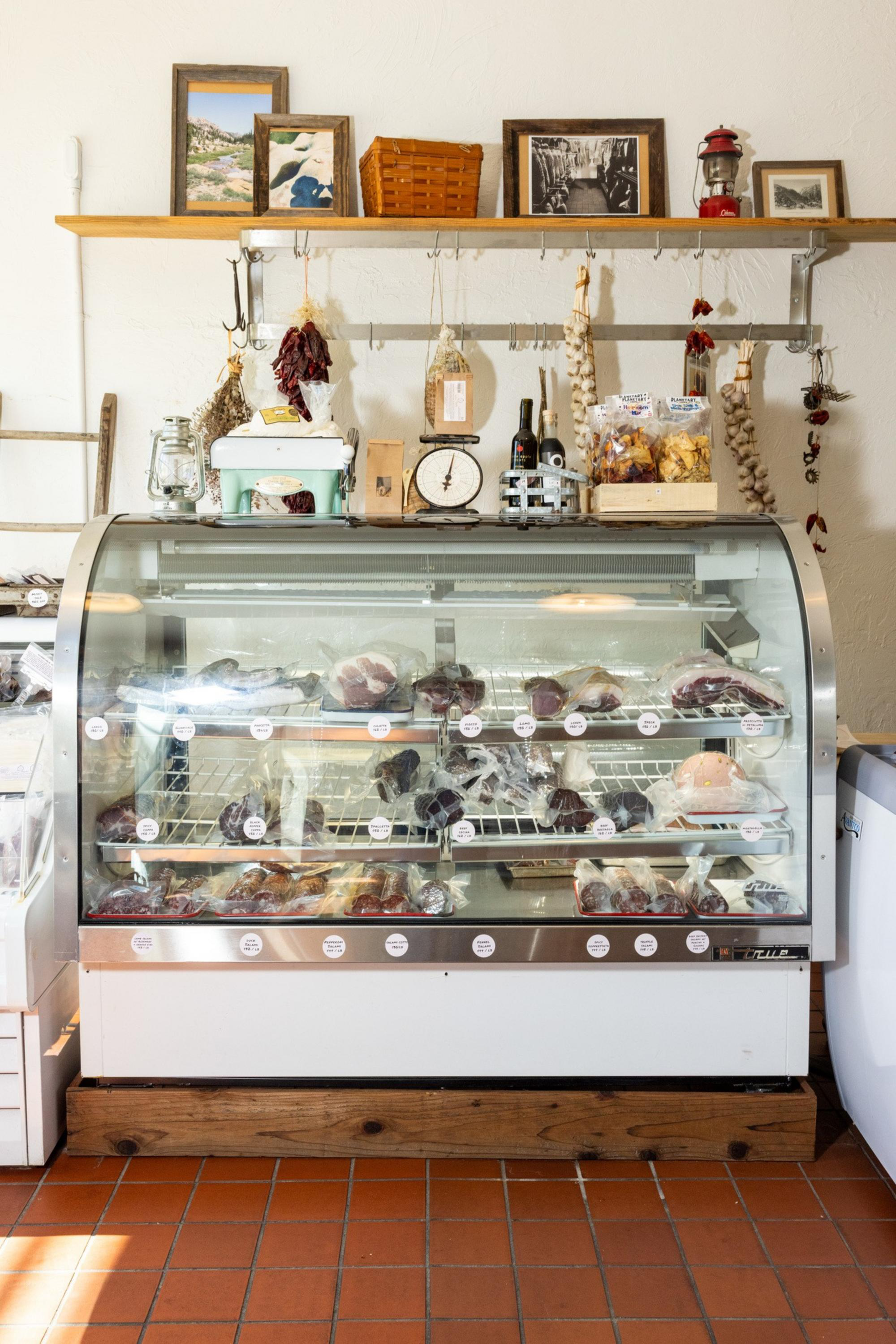 A refrigerated display case holds various packaged meats, with shelves above featuring framed pictures, hanging dried herbs, a scale, and a basket.