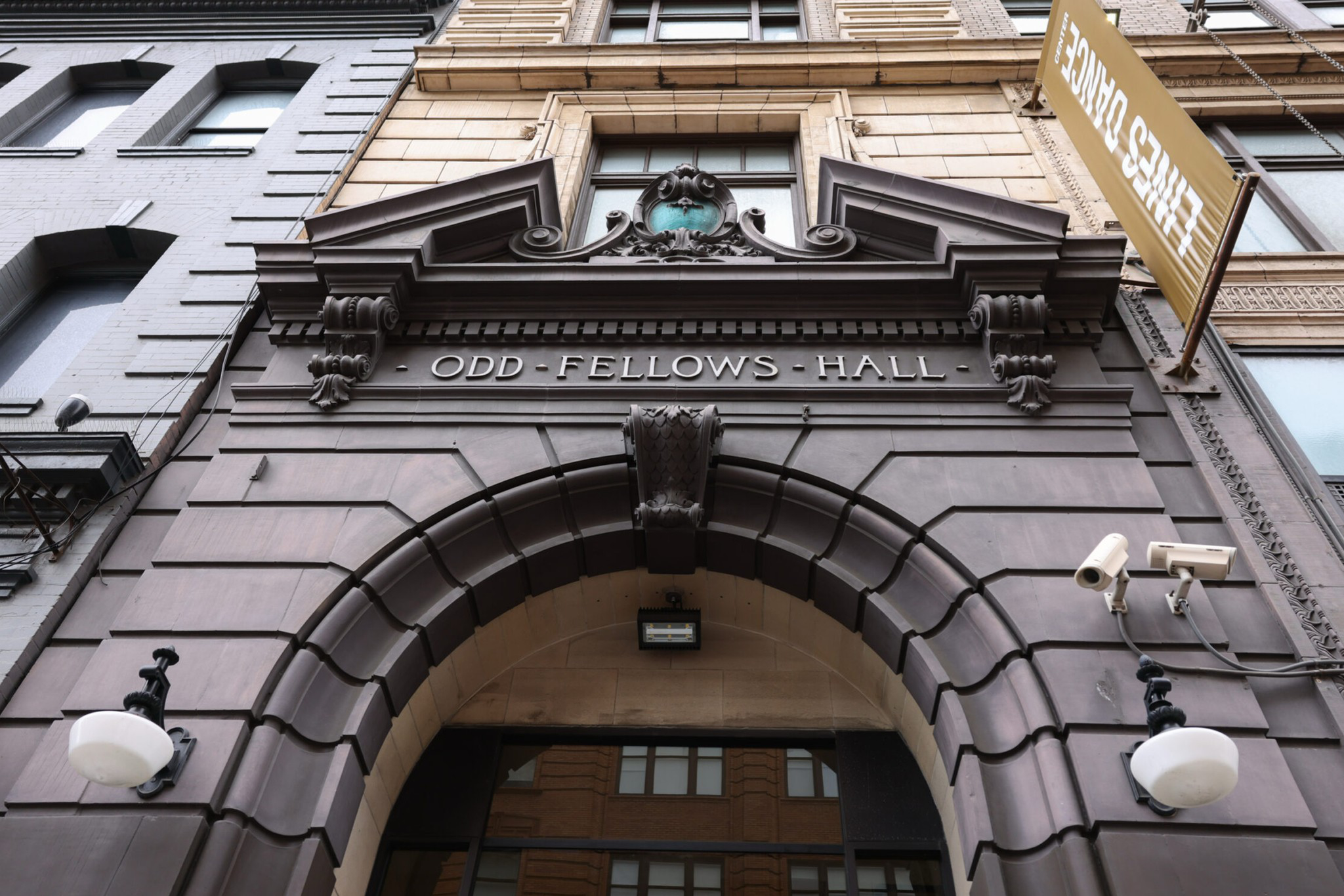 A stone archway with intricate details has the inscription "ODD FELLOWS HALL" above it, flanked by lamps and surveillance cameras.