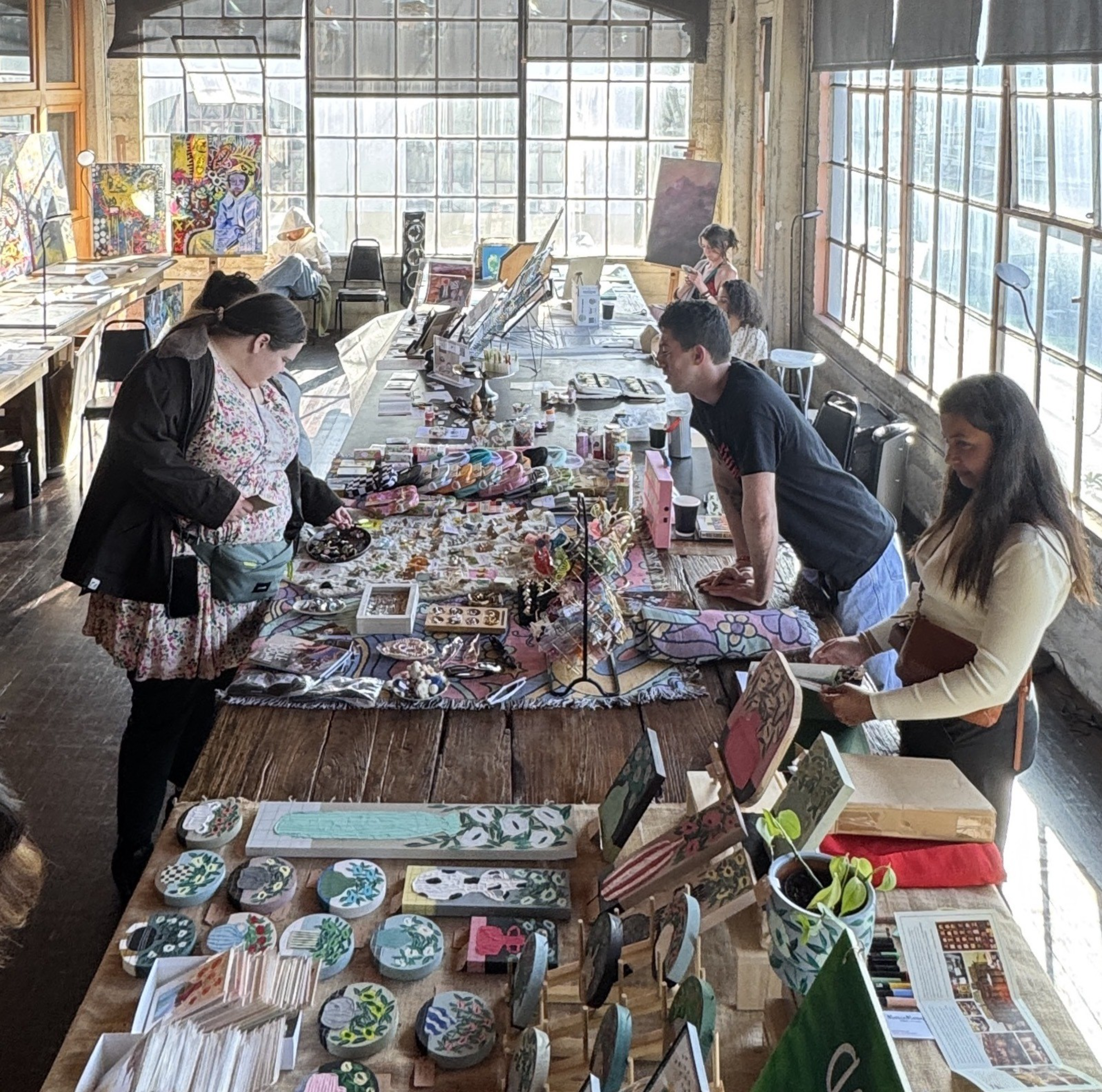 Three people examine colorful art pieces on a long table in a spacious, sunlit room with large windows and various paintings propped against the walls.