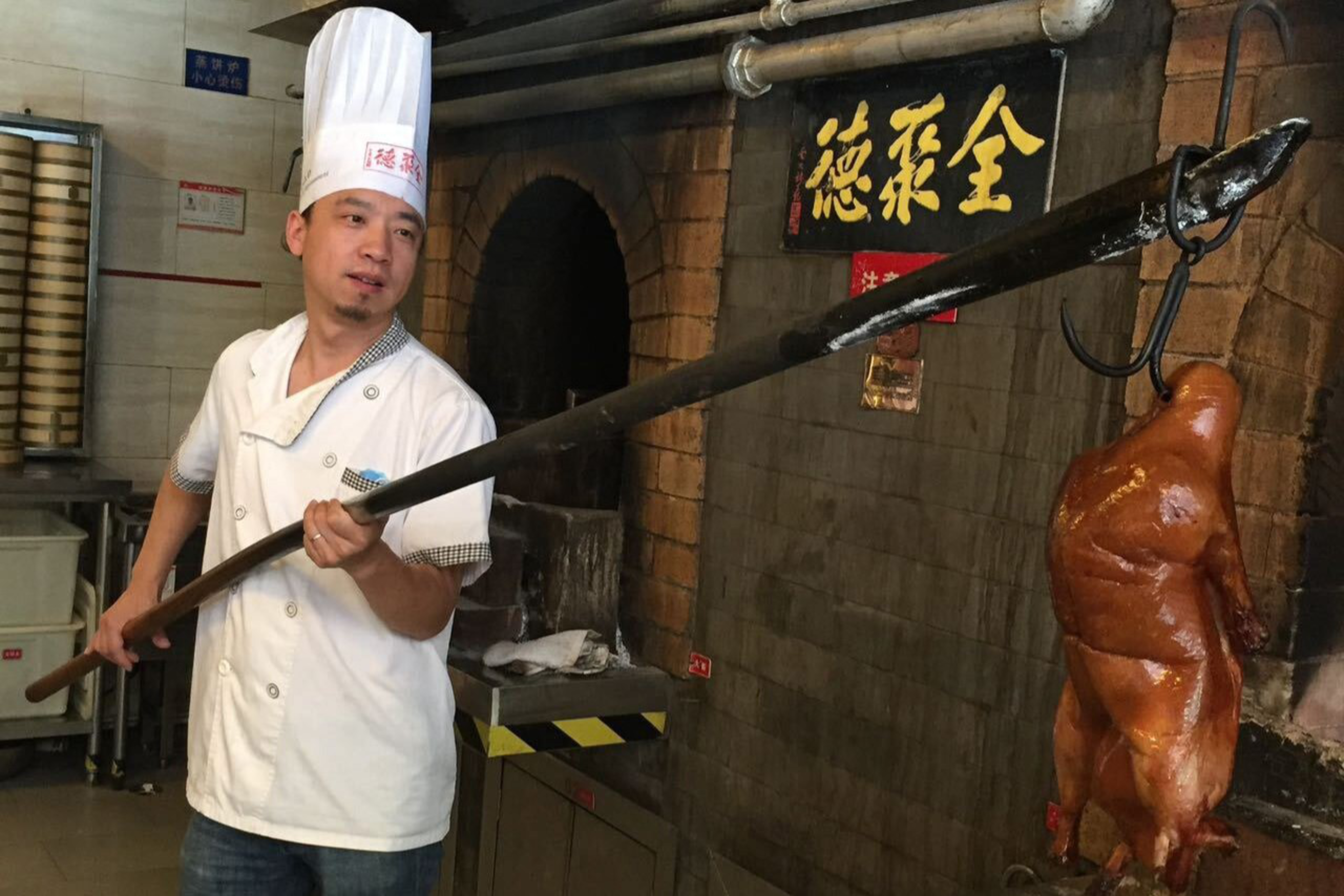 A chef in a white uniform and tall hat uses a long wooden pole to hang a roasted duck inside a brick oven.