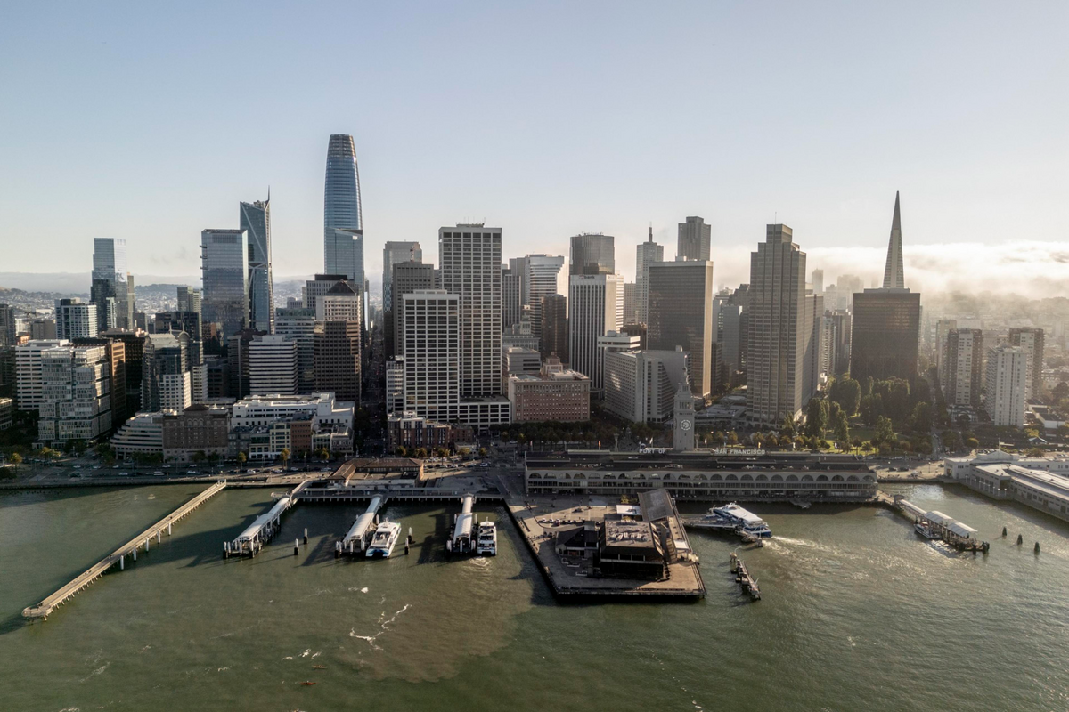A waterfront city skyline with multiple high-rise buildings, piers extending into the water, and boats docked along the shore.