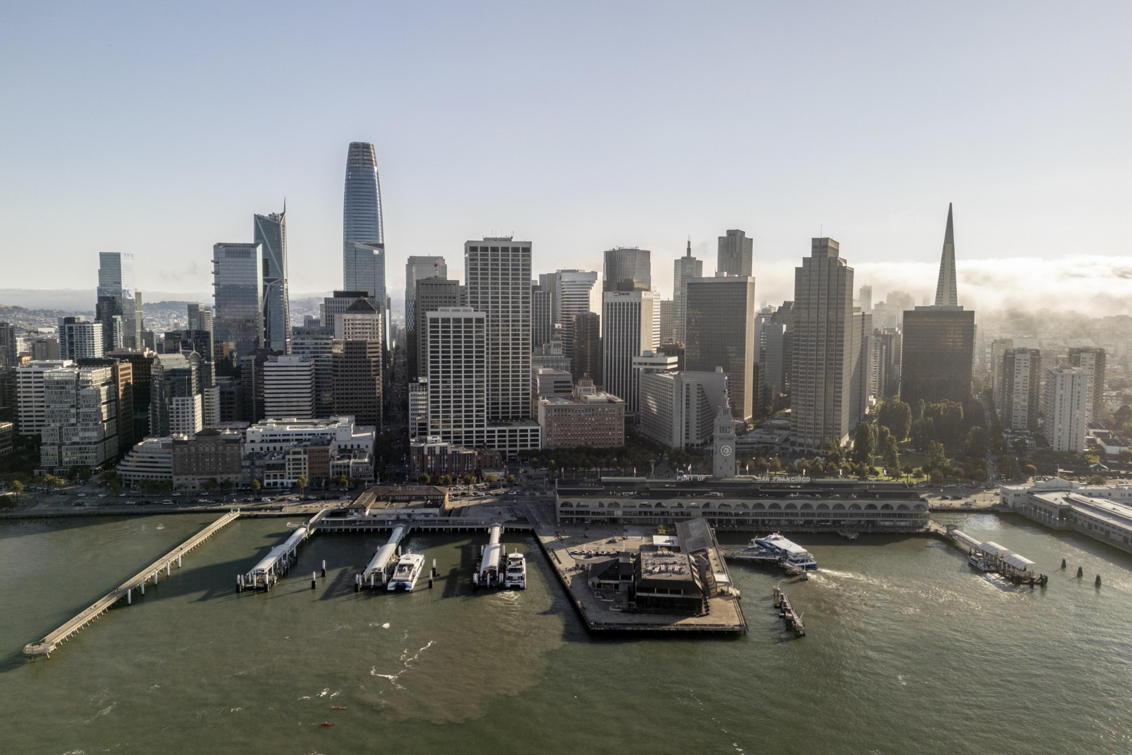 A waterfront city skyline with multiple high-rise buildings, piers extending into the water, and boats docked along the shore.