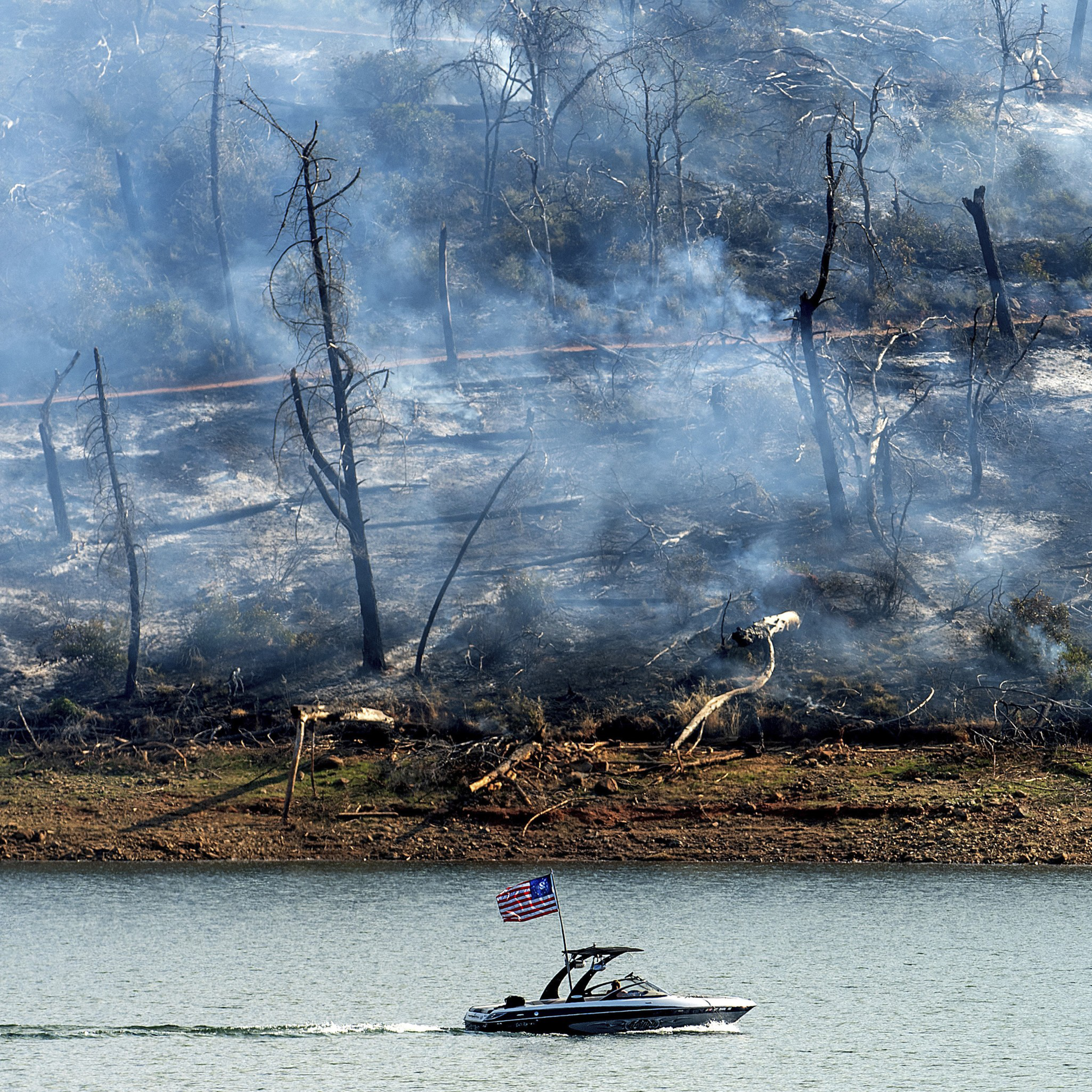 A motorboat with an American flag cruises on a lake, while a smoky, charred, and barren forest is visible on the opposite shore, indicating a recent fire.