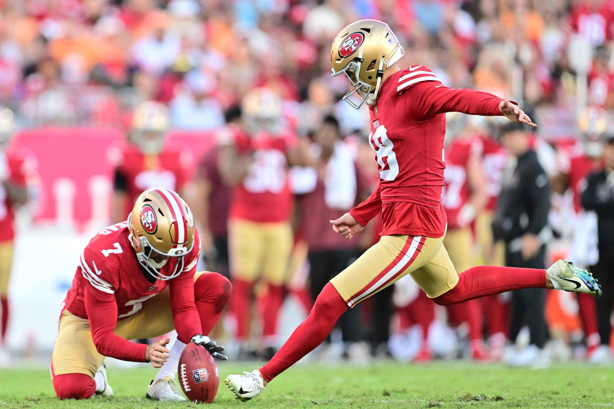 A football player in a red and gold uniform kicks the ball while another teammate holds it steady on a grassy field.
