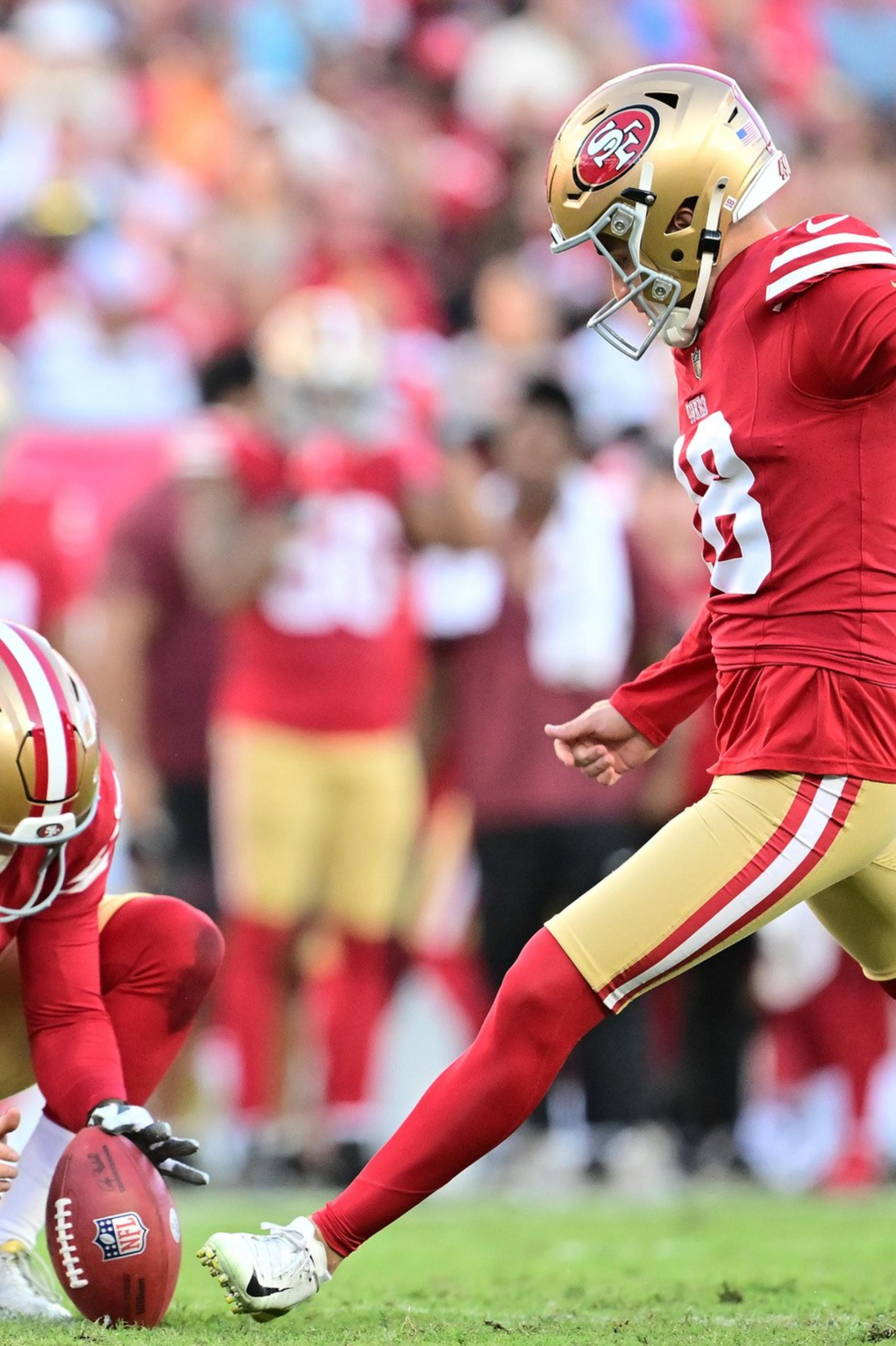 A football player in a red and gold uniform kicks the ball while another teammate holds it steady on a grassy field.