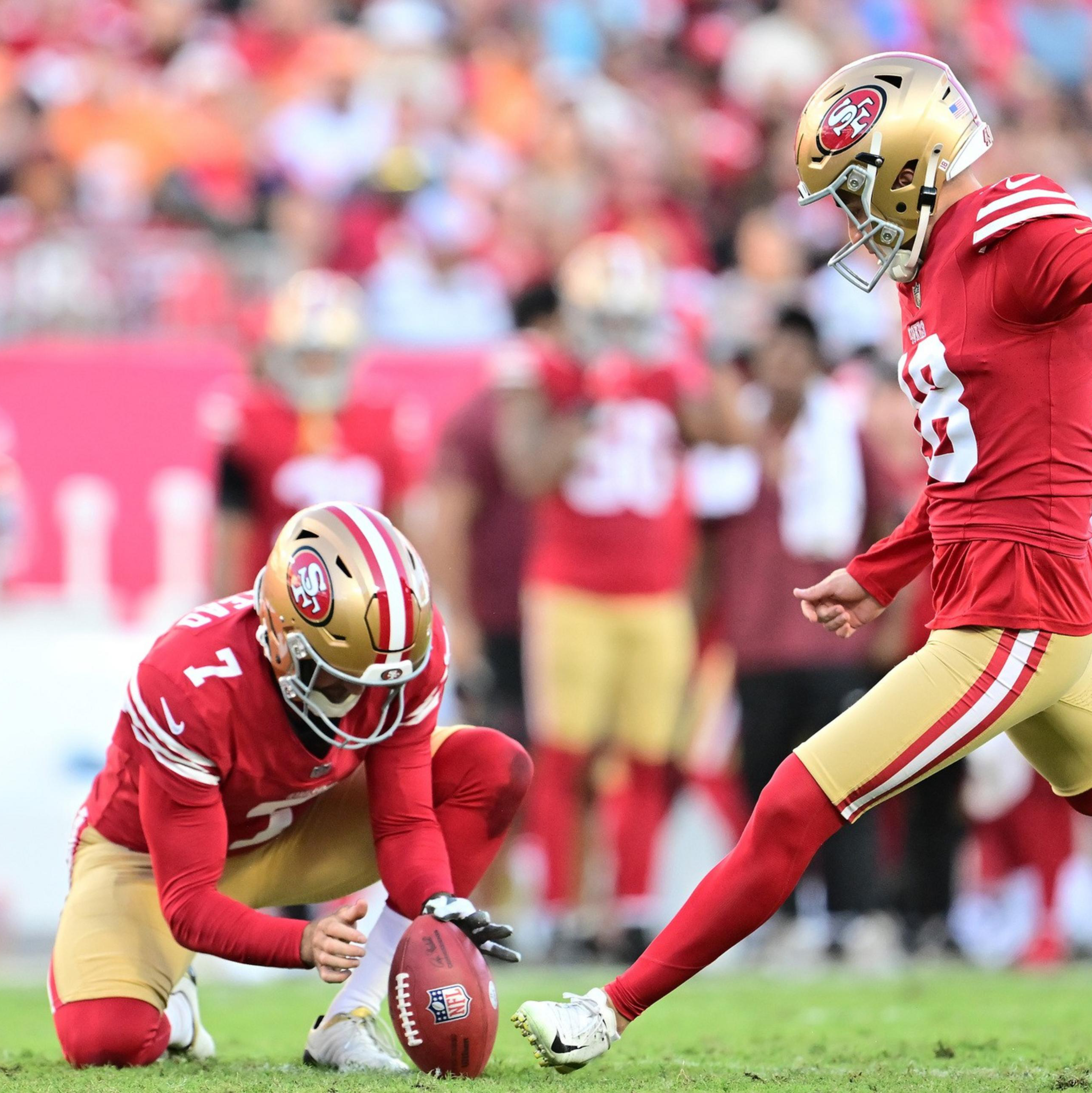 A football player in a red and gold uniform kicks the ball while another teammate holds it steady on a grassy field.