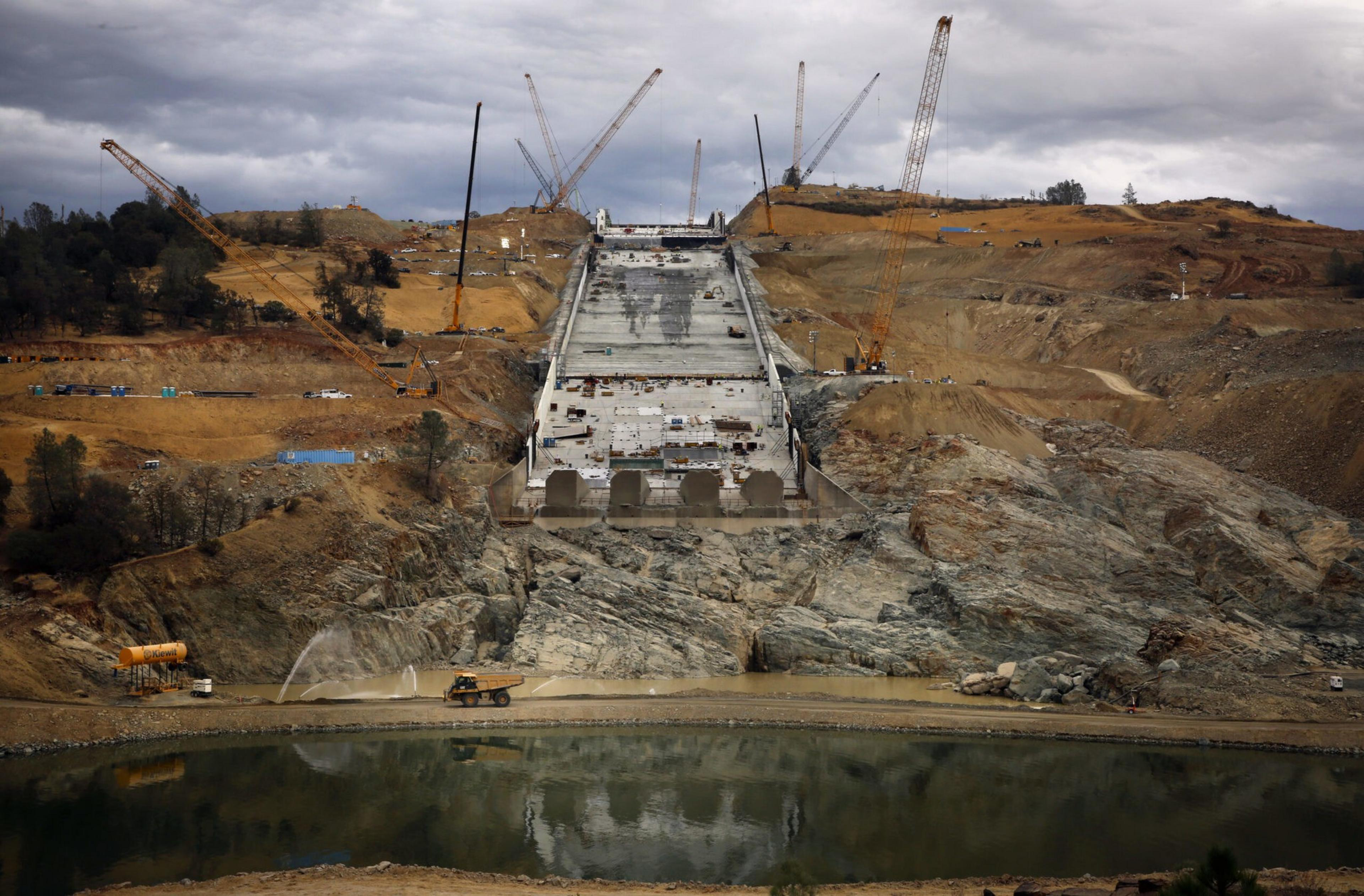 A large dam under construction on a rocky hillside with cranes and construction vehicles around, and a body of water at the base.