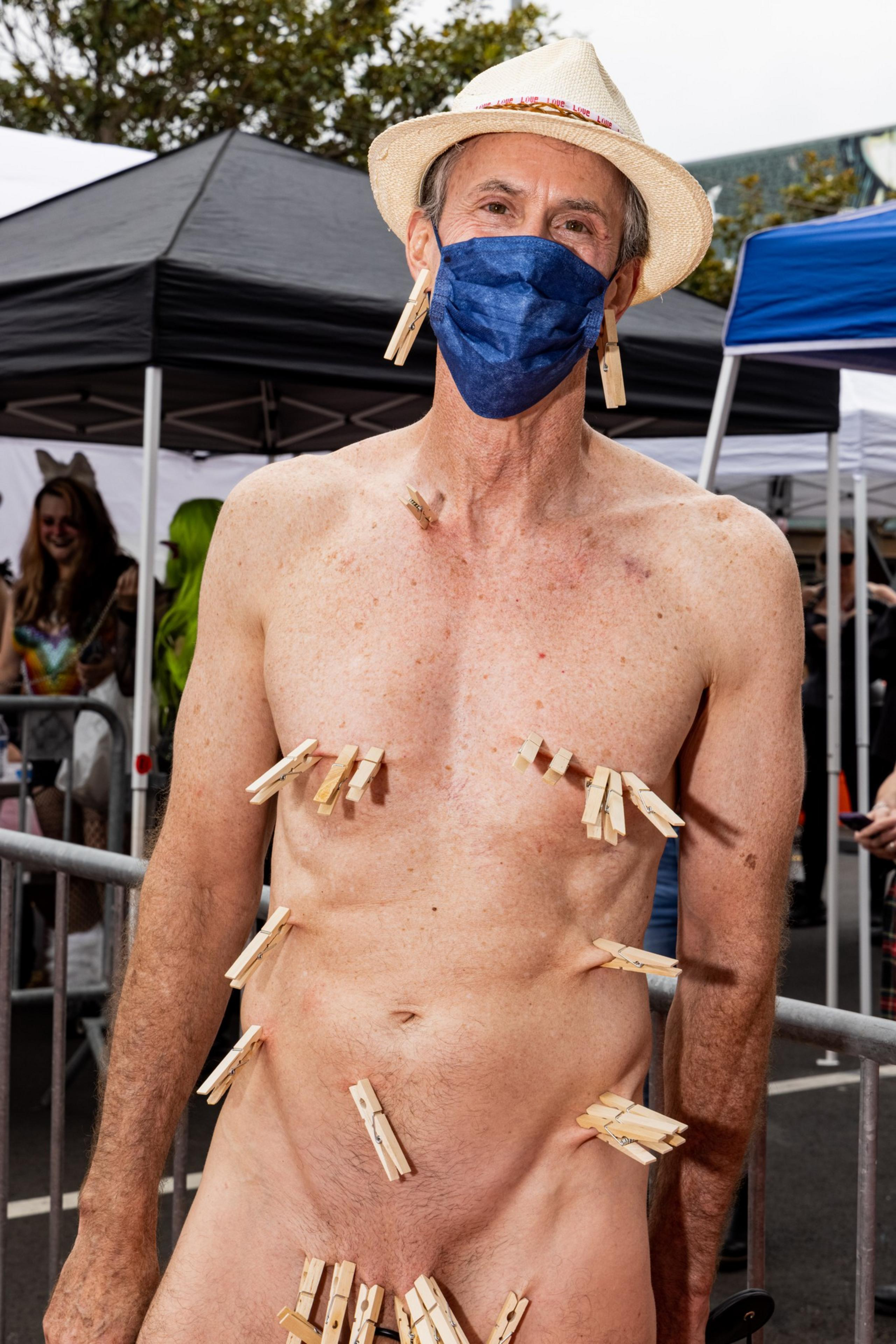 A shirtless man wearing a straw hat and blue face mask has multiple wooden clothespins clipped to his chest, torso, and groin area.