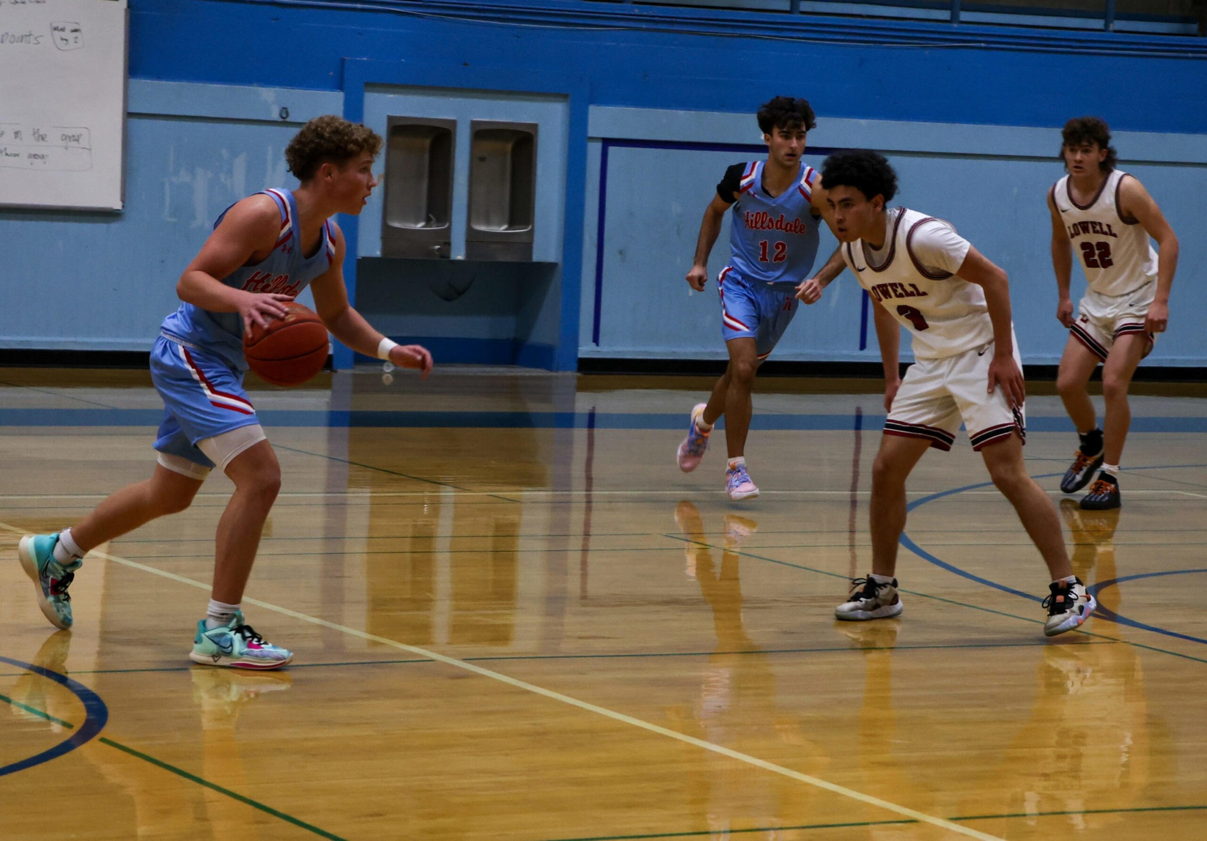 A basketball player in a blue uniform dribbles the ball while two opponents in white uniforms defend and another blue-uniformed player approaches.