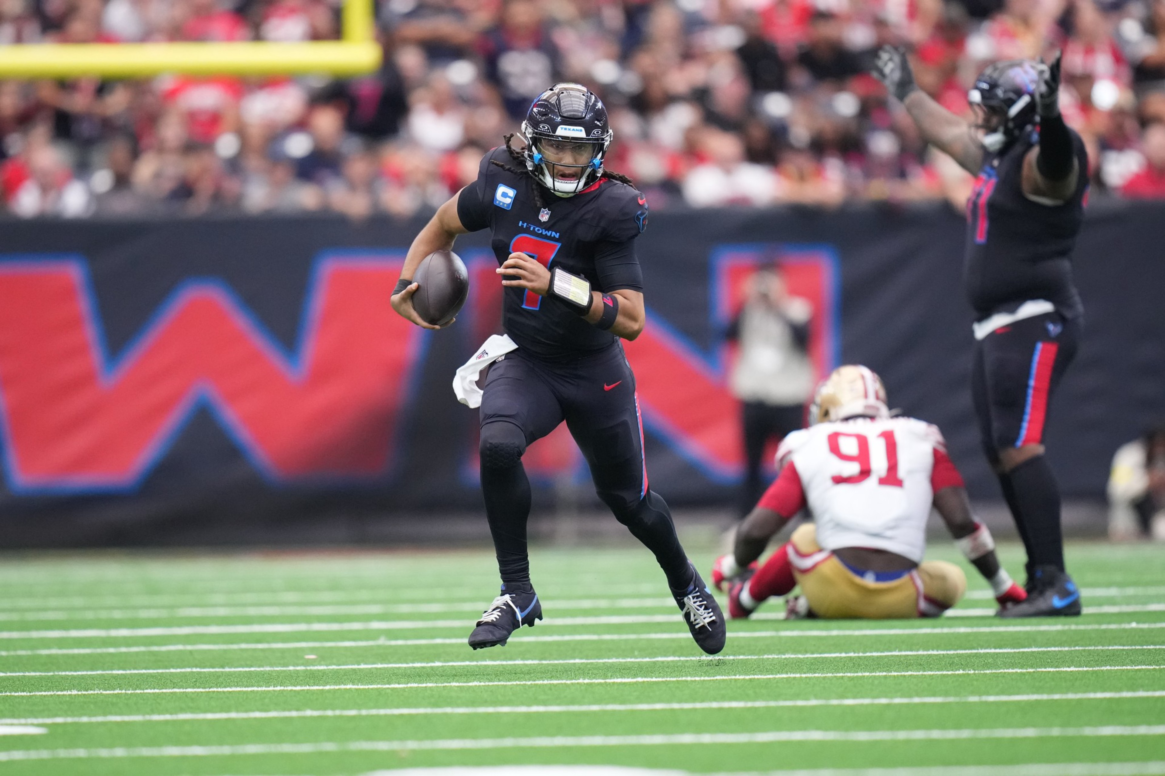 A football player in a black uniform runs with the ball as a player in a white and red uniform sits on the field behind him and another in black raises his arms.