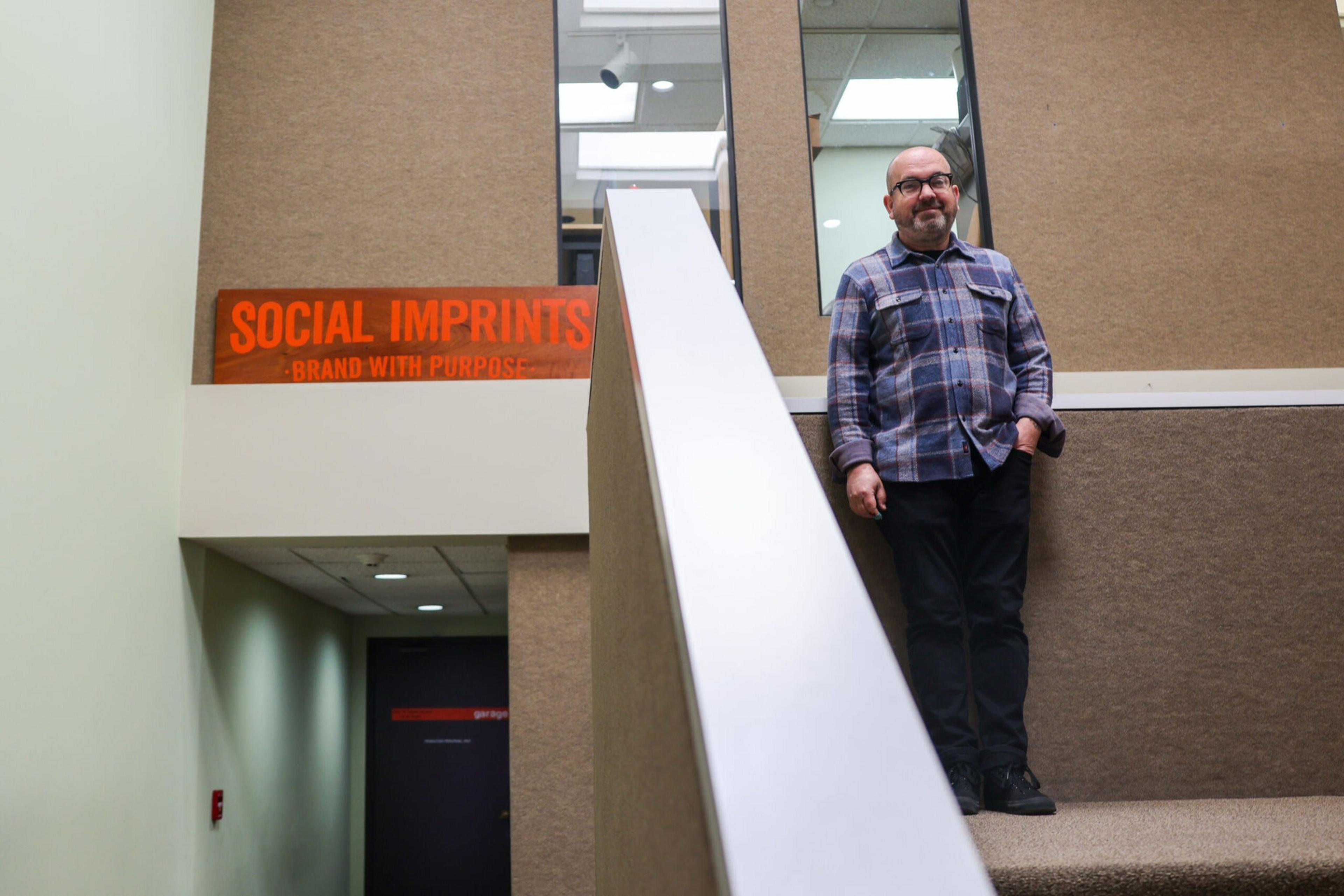 A man with glasses and a plaid shirt stands on a carpeted staircase next to a sign that reads "SOCIAL IMPRINTS - BRAND WITH PURPOSE."