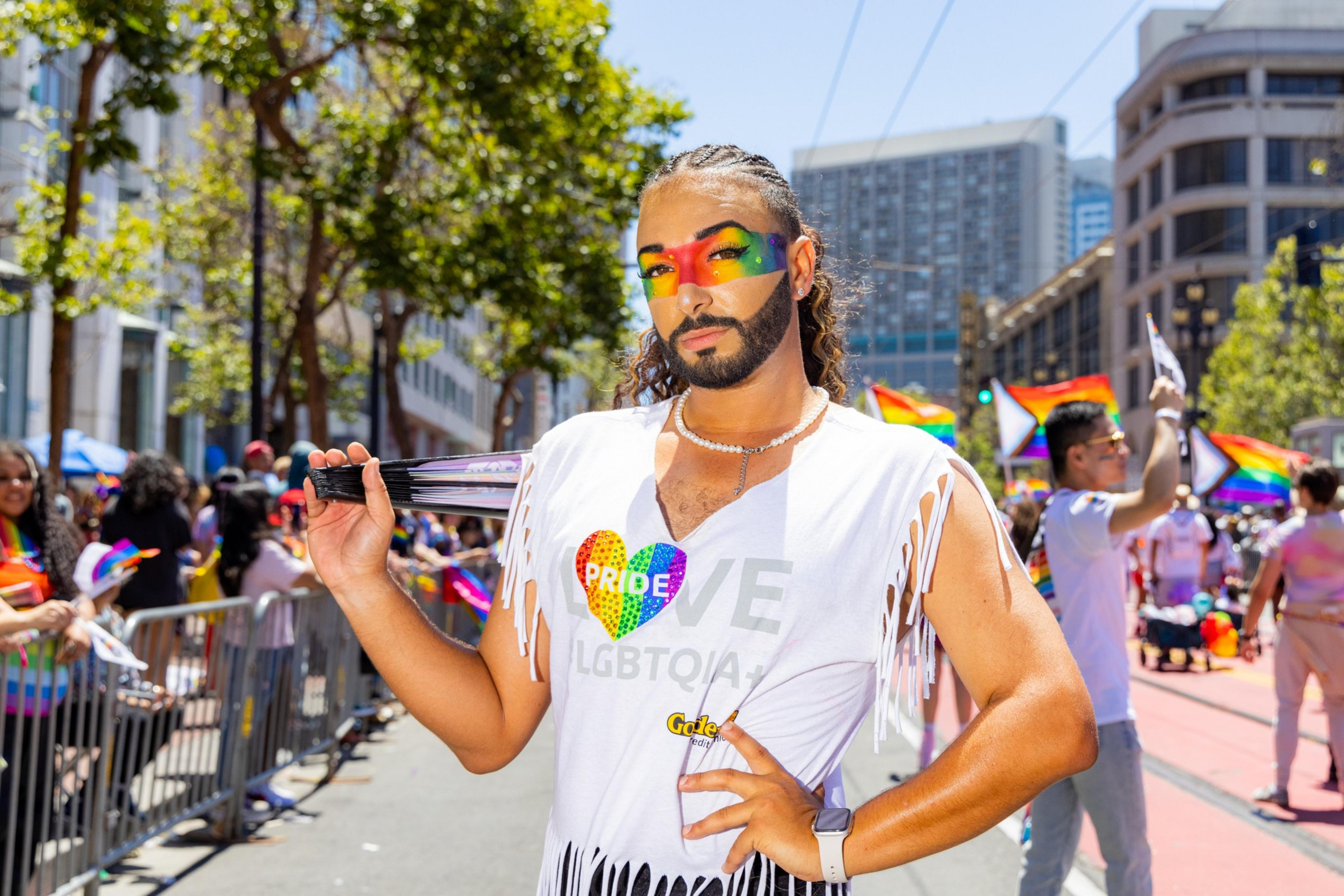 A person with rainbow face paint and a fringed white shirt that says “PRIDE” stands outdoors, holding a fan, among a crowd with rainbow flags and trees in the background.