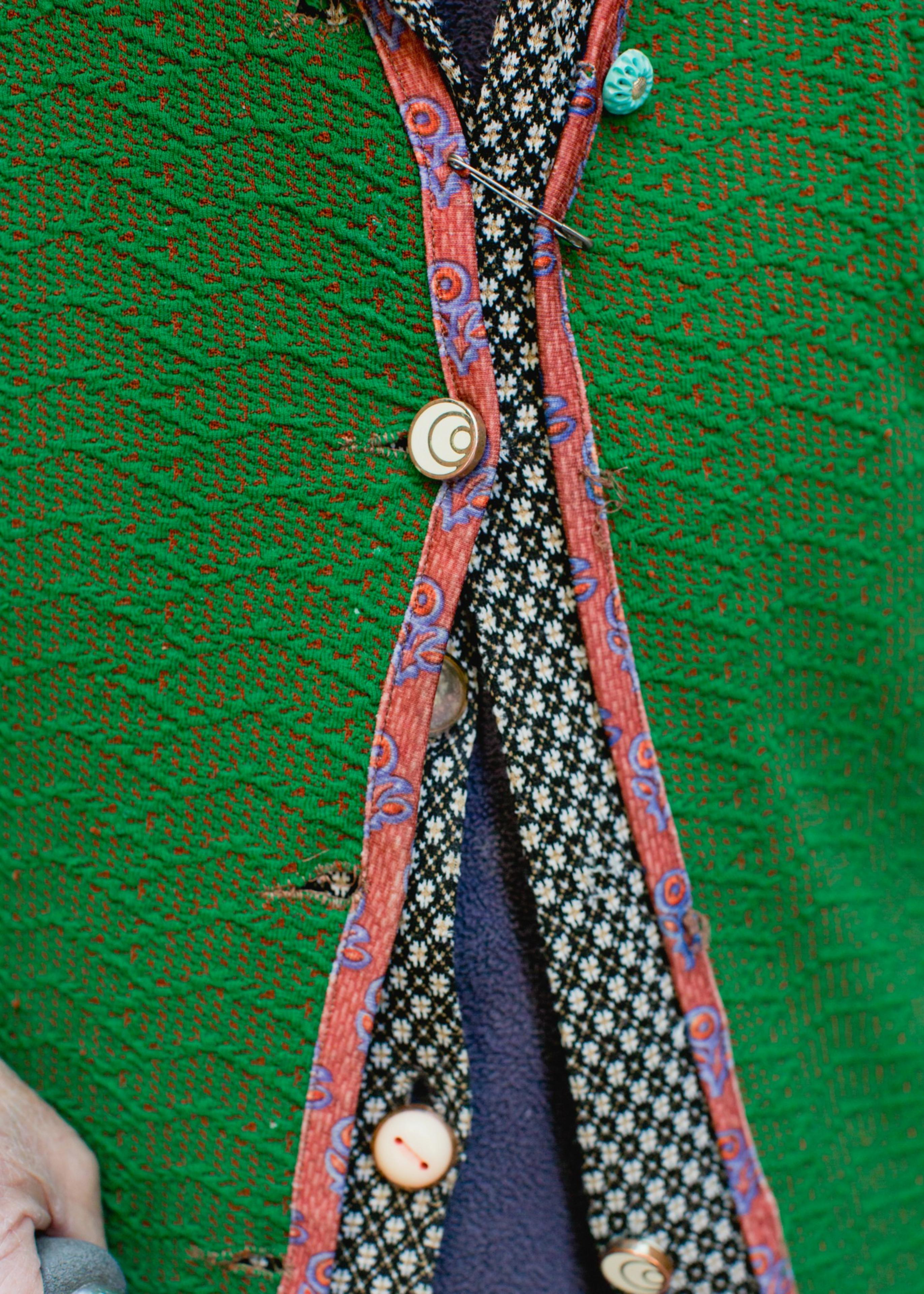 A green knit garment with a red floral trim, fastened with mismatched buttons and a safety pin, reveals a black and white patterned shirt underneath.