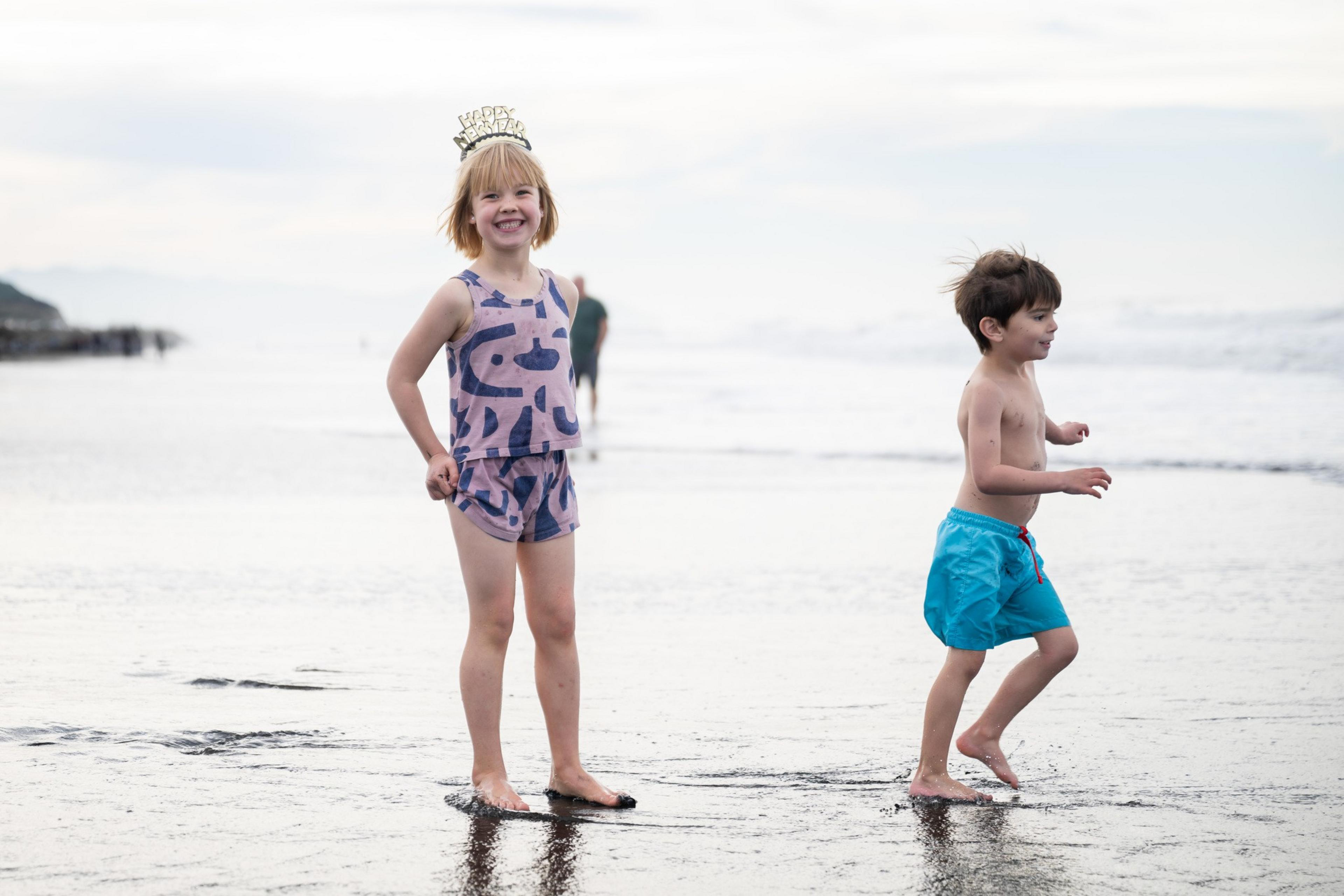 Two children are playing on a beach. One, wearing a crown and matching outfit, smiles at the camera. The other, in blue shorts, runs joyfully.