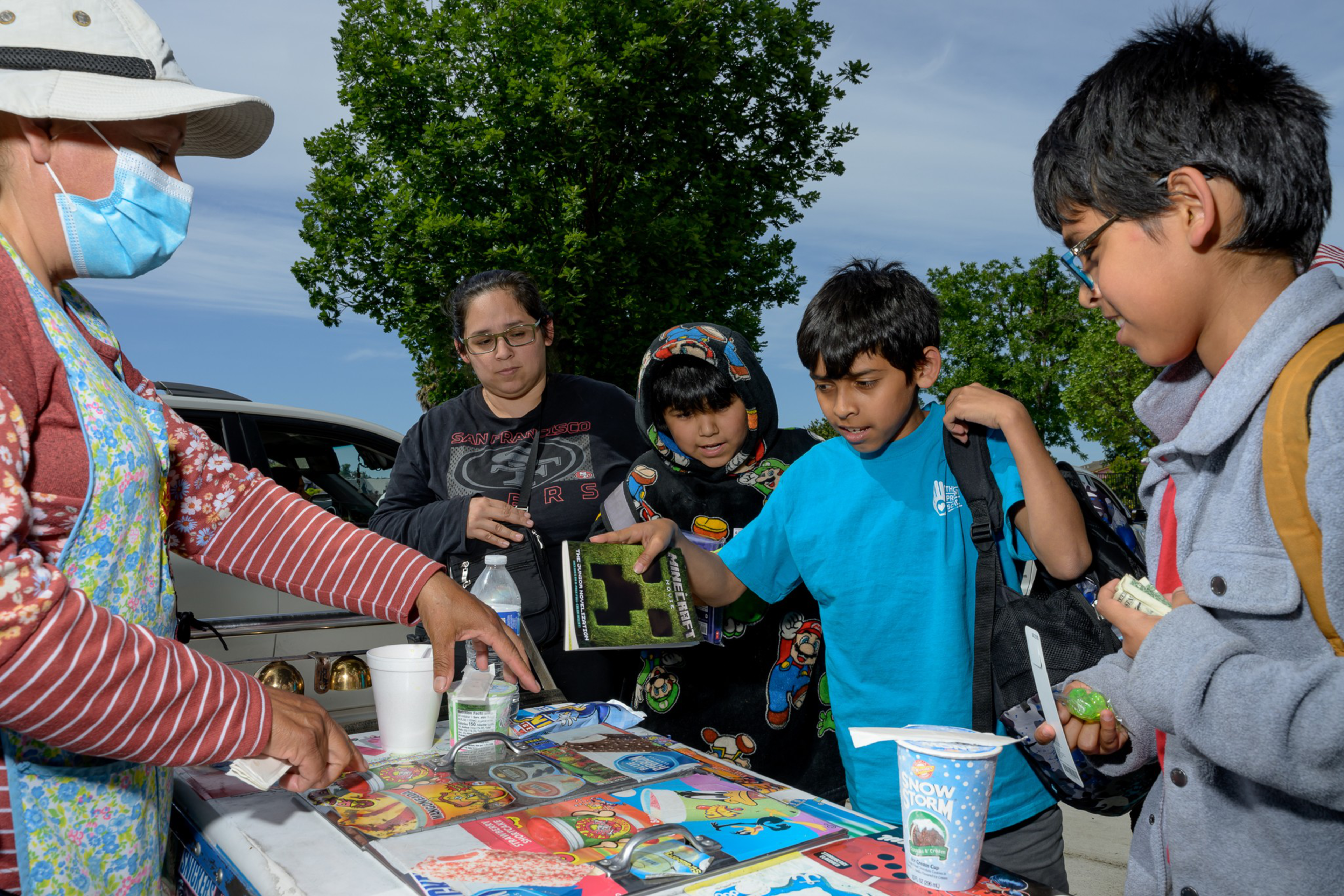 A vendor wearing a mask serves three children and a woman outdoors. The table has snacks, toys, and a book; one child holds a Minecraft book.