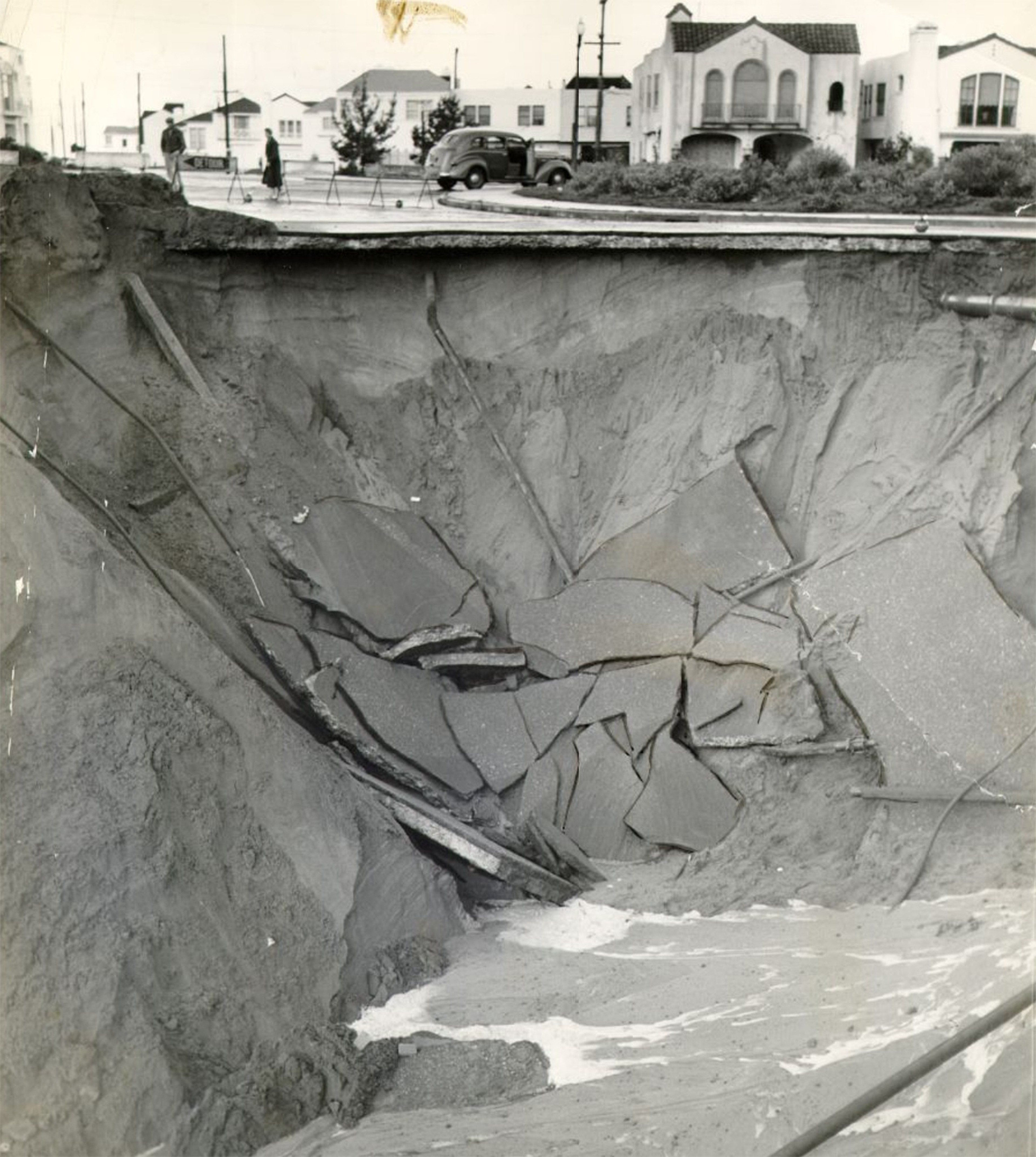 A large sinkhole has collapsed a section of a paved road, exposing broken asphalt slabs and earth beneath, with houses and a car in the background.