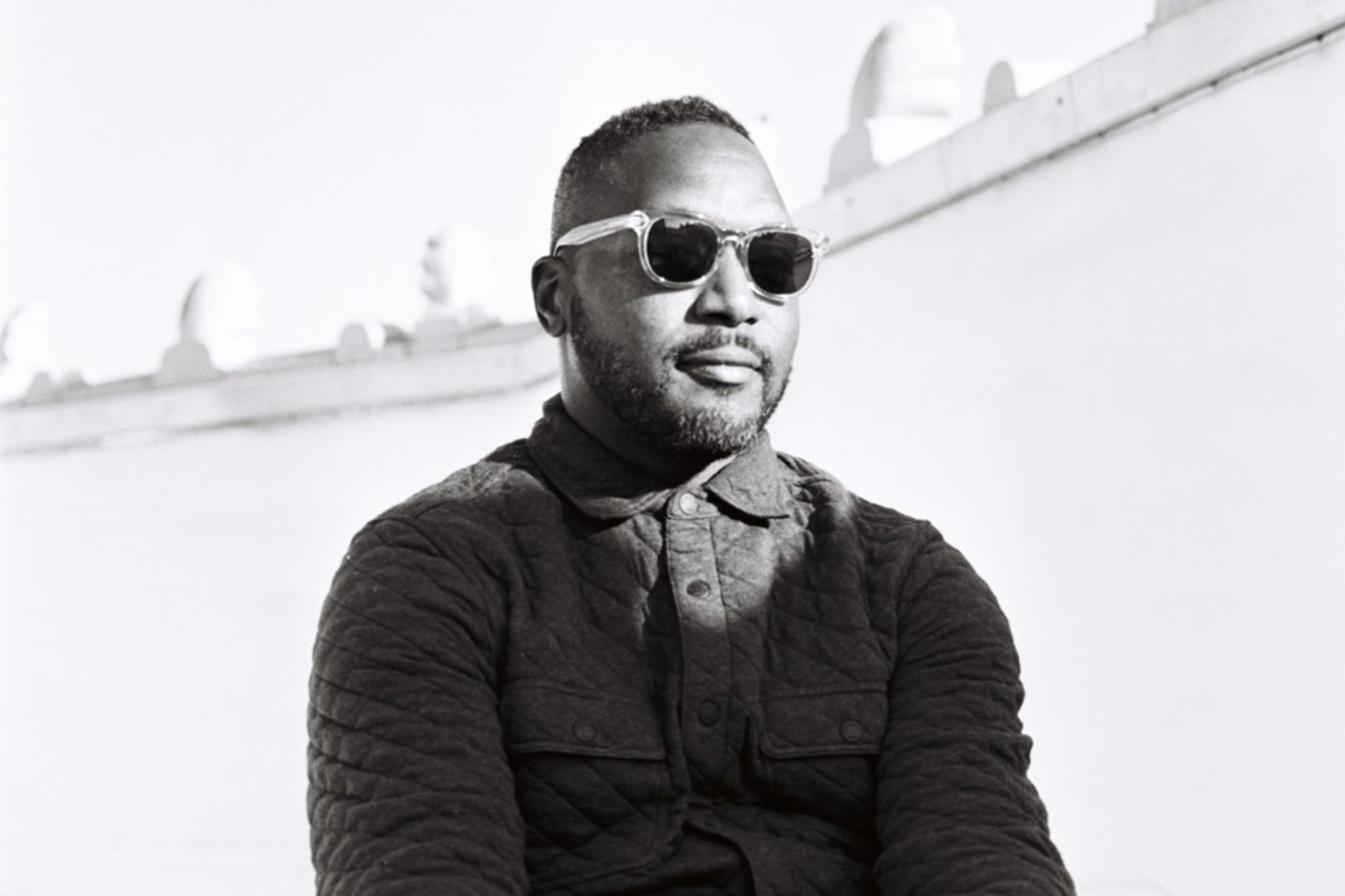 A man with a short beard and sunglasses sits against a plain white wall, wearing a textured, buttoned jacket and looking slightly to the side.