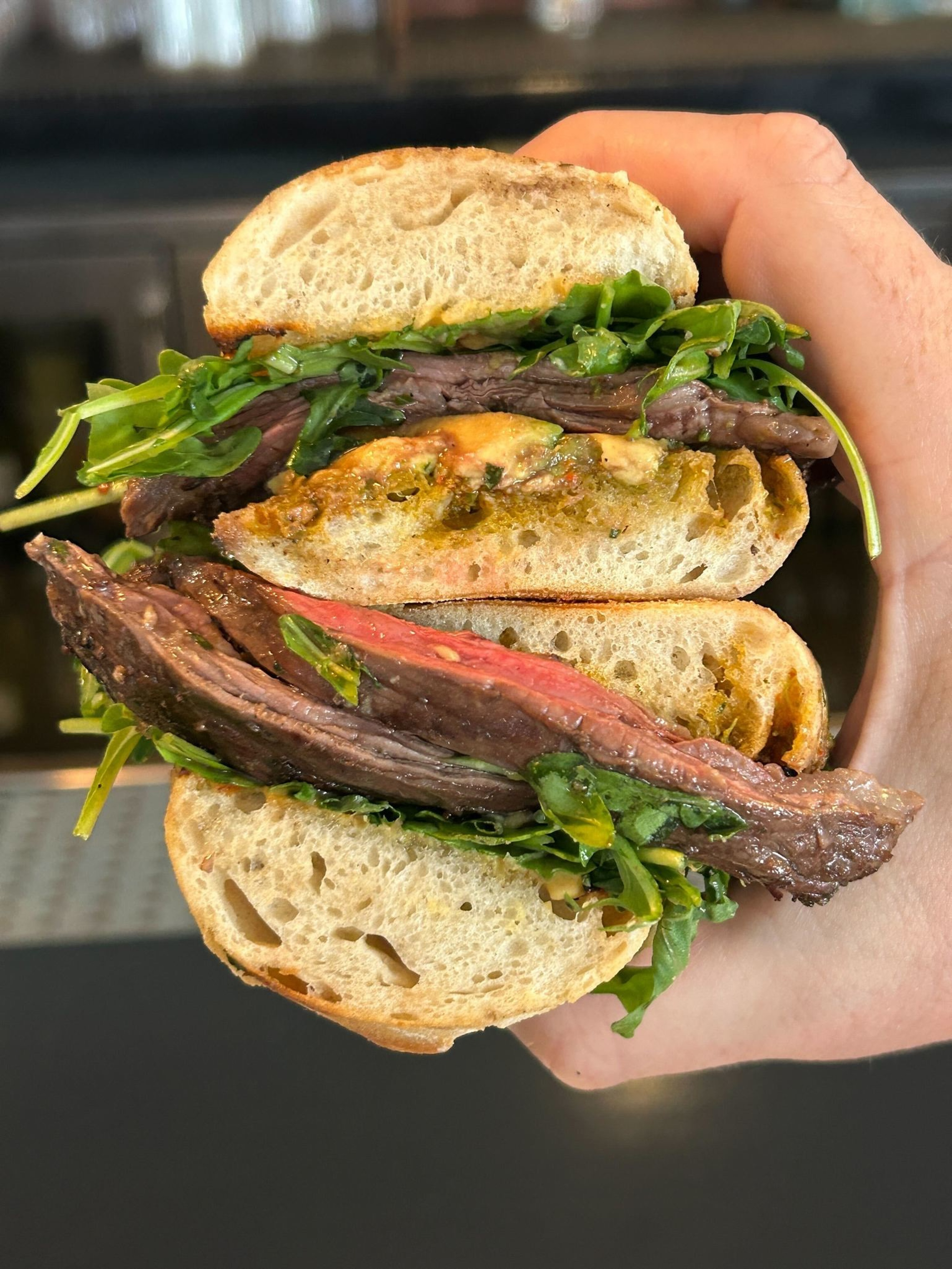 A hand holds a sandwich with medium-rare steak, fresh green leafy vegetables, and a thick slice of crusty bread.