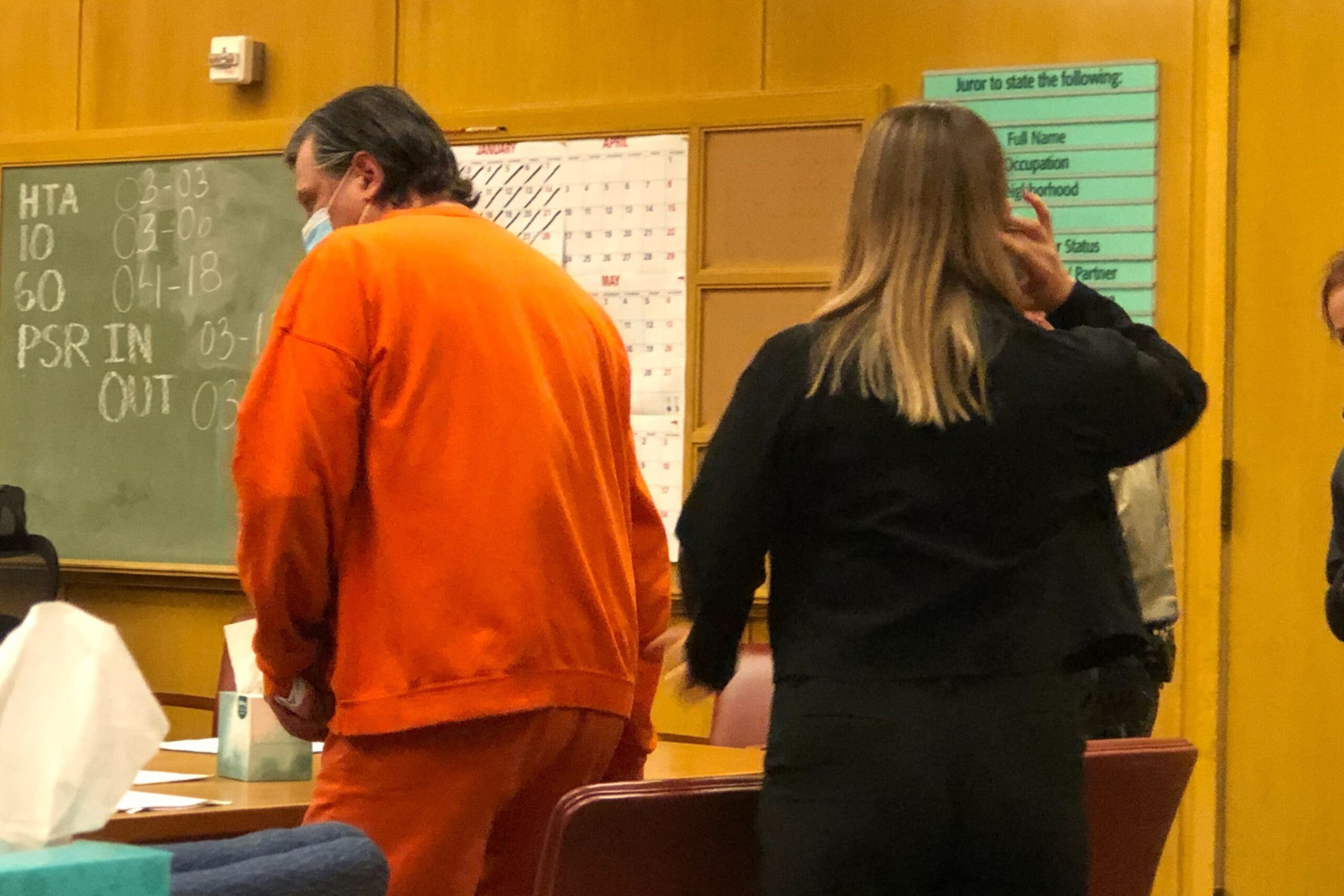 A man in an orange prison jumpsuit and a woman in black stand in a wood-paneled room with a chalkboard, calendar, and jury instructions poster.