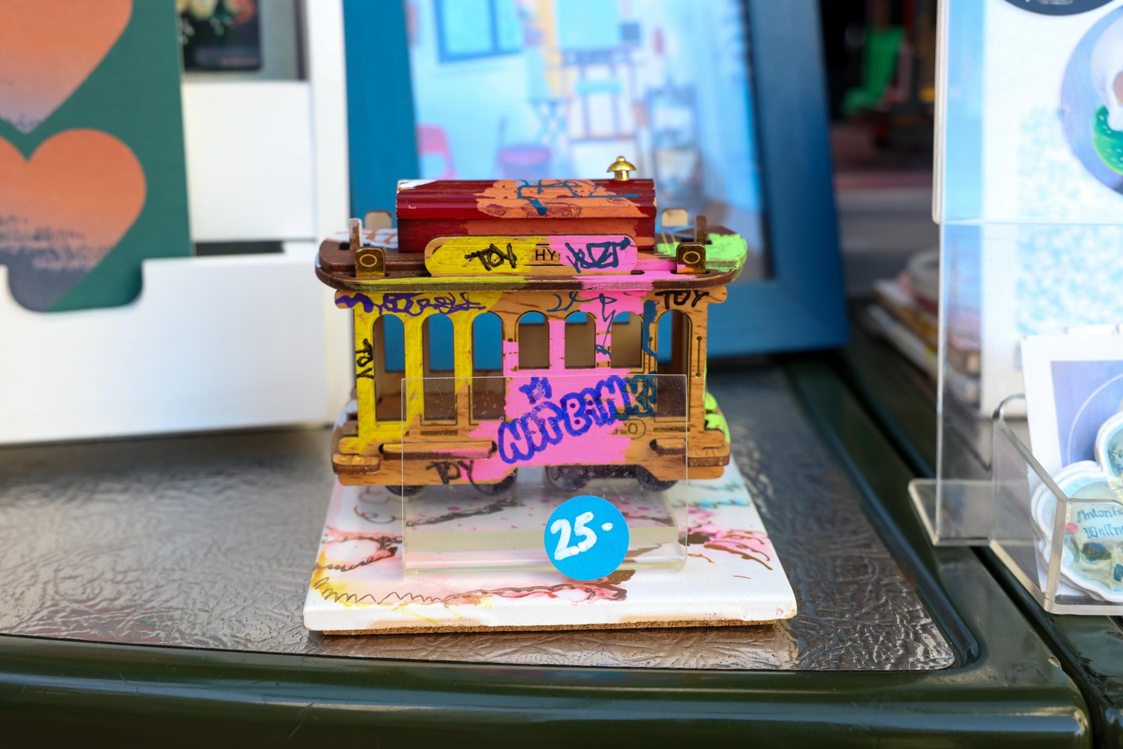 A small, colorful wooden cable car model with graffiti-style paint rests on a stand marked with a blue "25.-" price sticker.