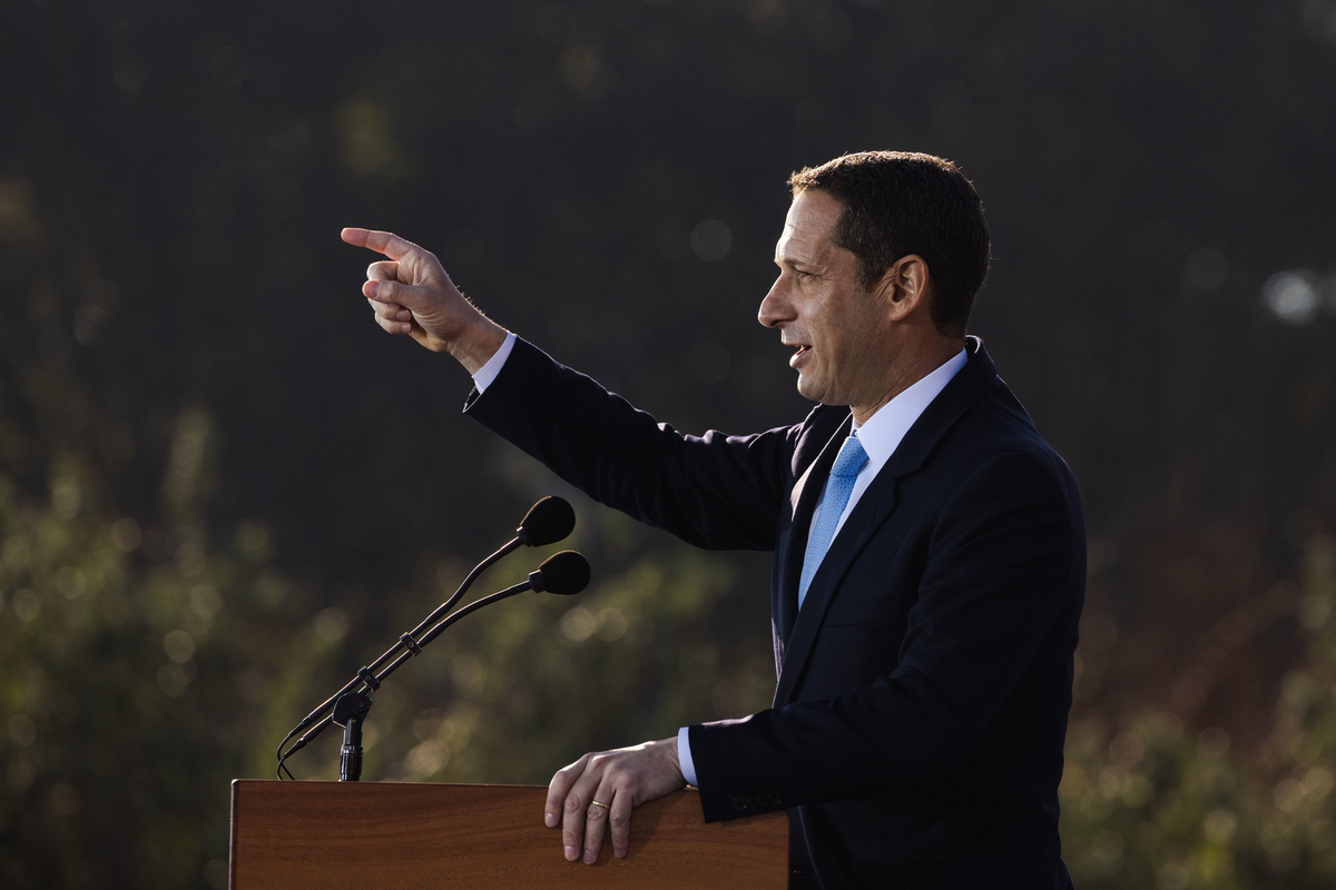 A man in a suit and blue tie stands at a podium with two microphones, pointing forward while speaking outdoors with a blurred natural background.