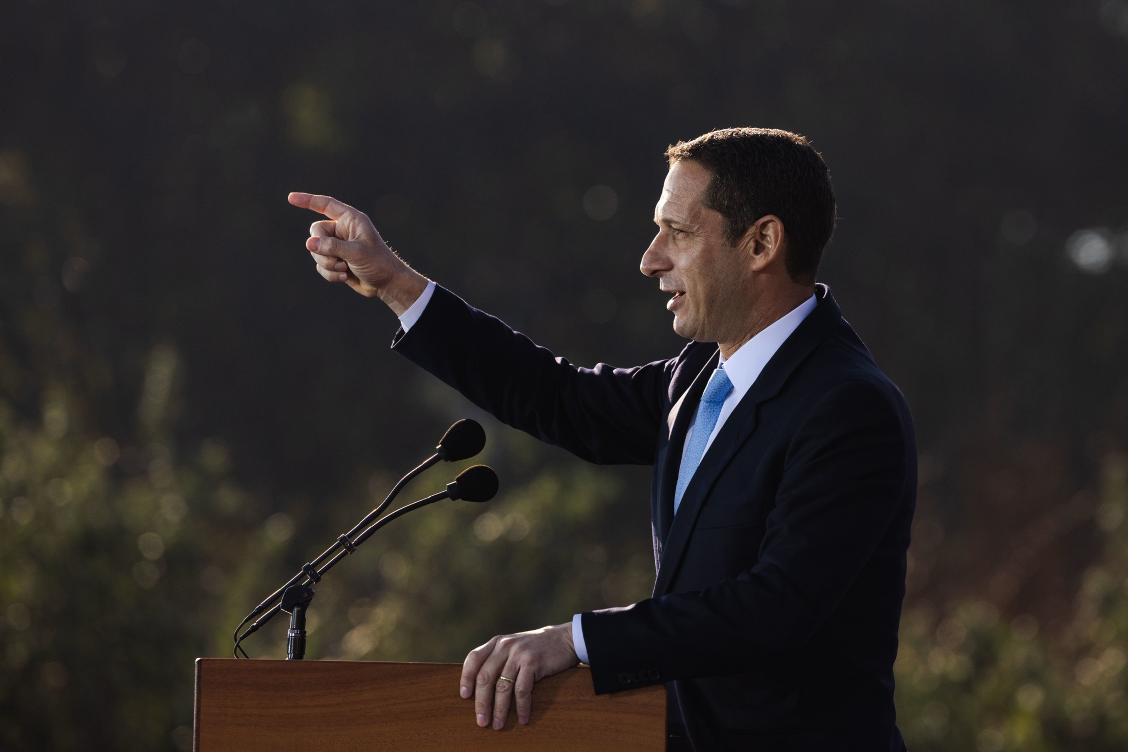 A man in a suit and blue tie stands at a podium with two microphones, pointing forward while speaking outdoors with a blurred natural background.