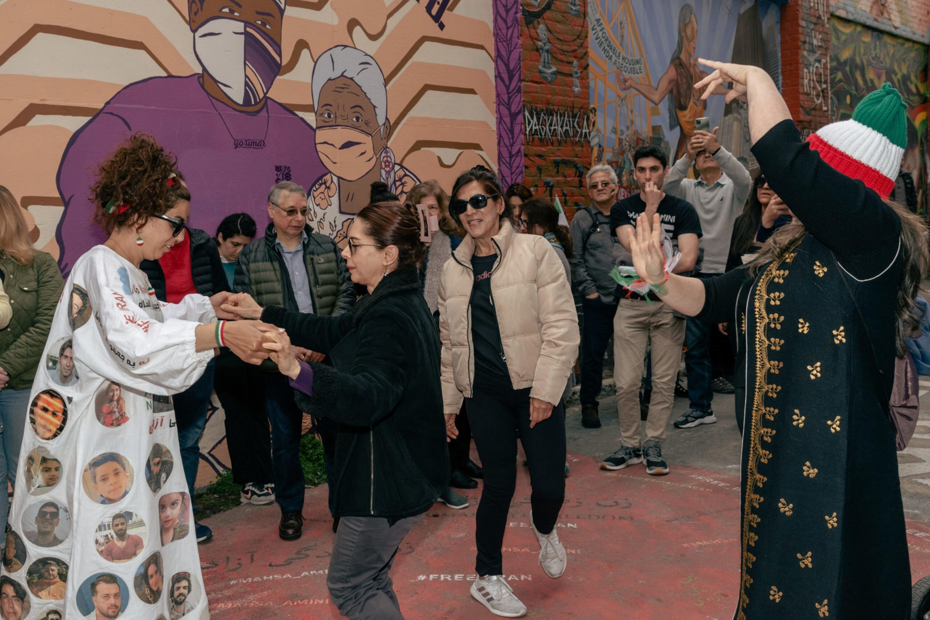 A group of people dance and watch on a street with colorful murals depicting masked faces and figures in the background.