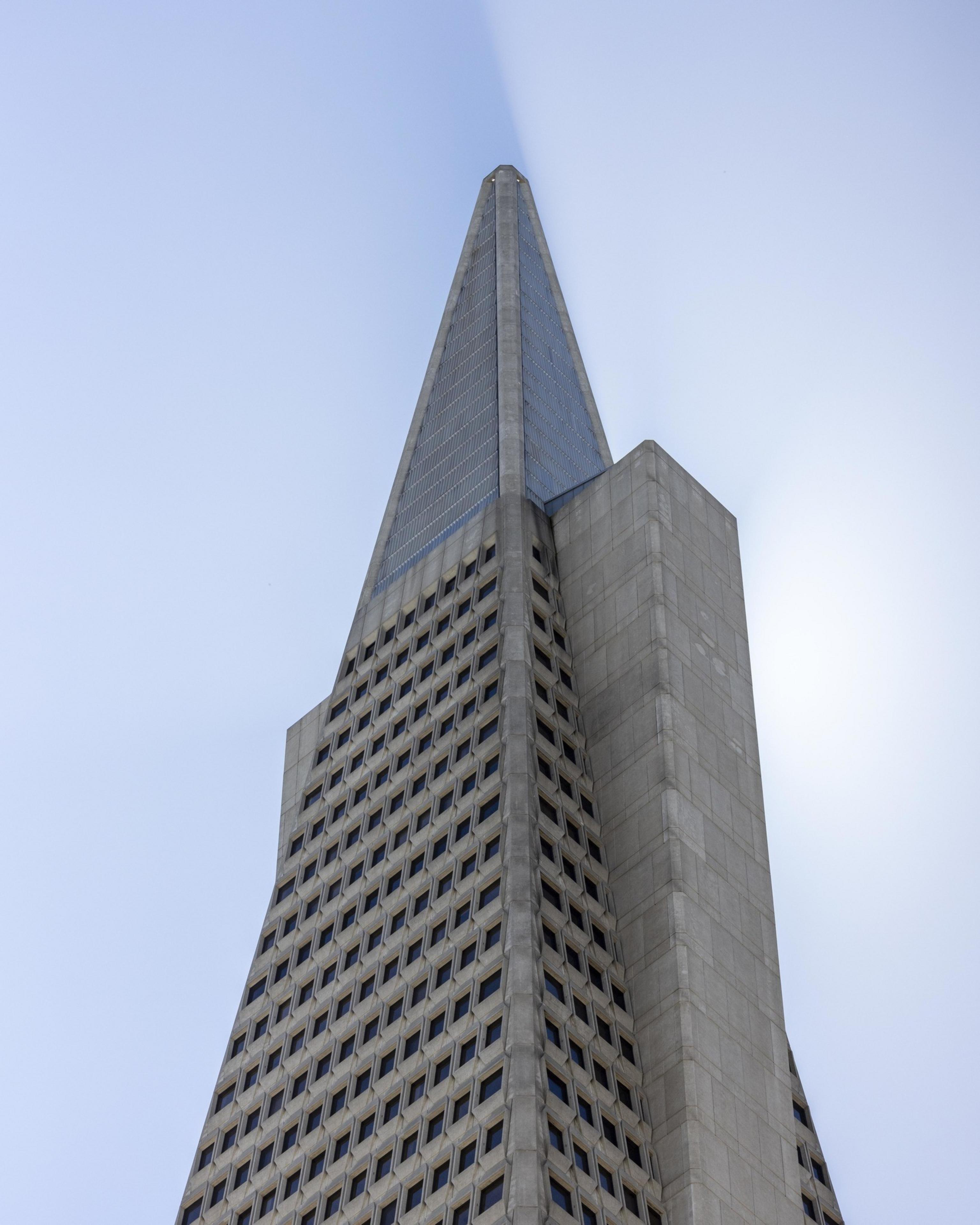 The image shows a tall, gray pyramidal building with a grid of small square windows, tapering to a sharp point at the top against a clear blue sky.