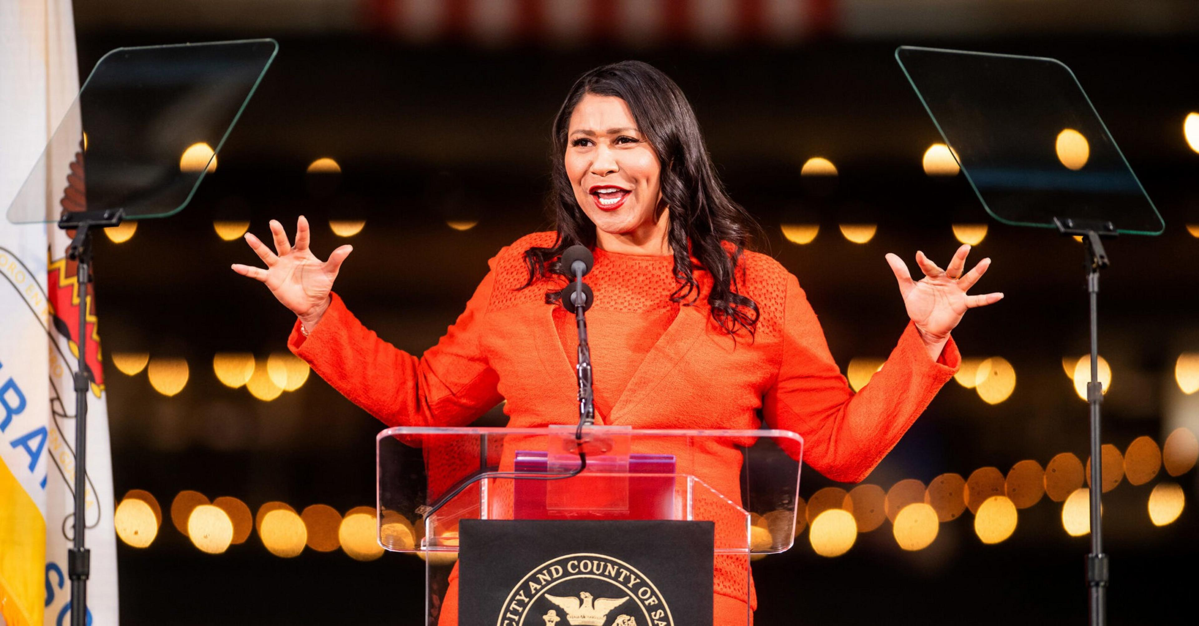 A woman in a red outfit speaks passionately behind a podium with a seal, gesturing widely with both hands raised. Two transparent teleprompters flank her.