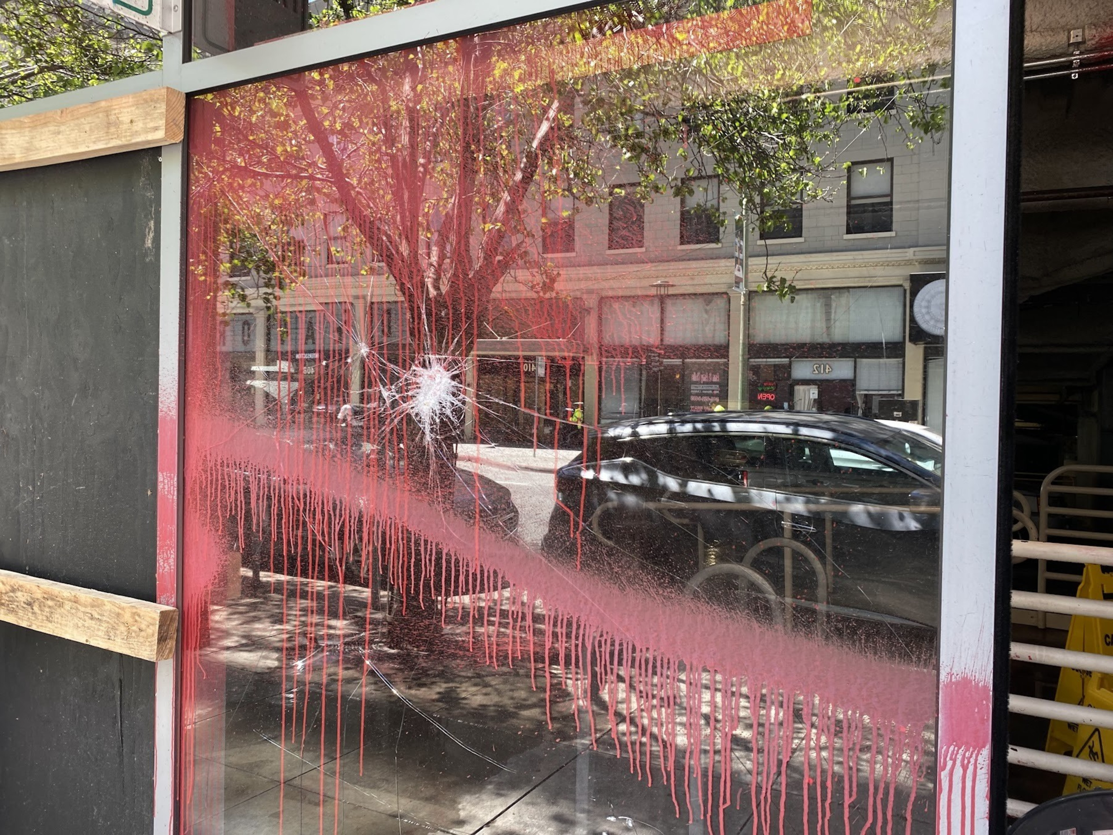 A shattered glass window with white and red spray paint drips, showing a reflection of urban buildings and a passing car.