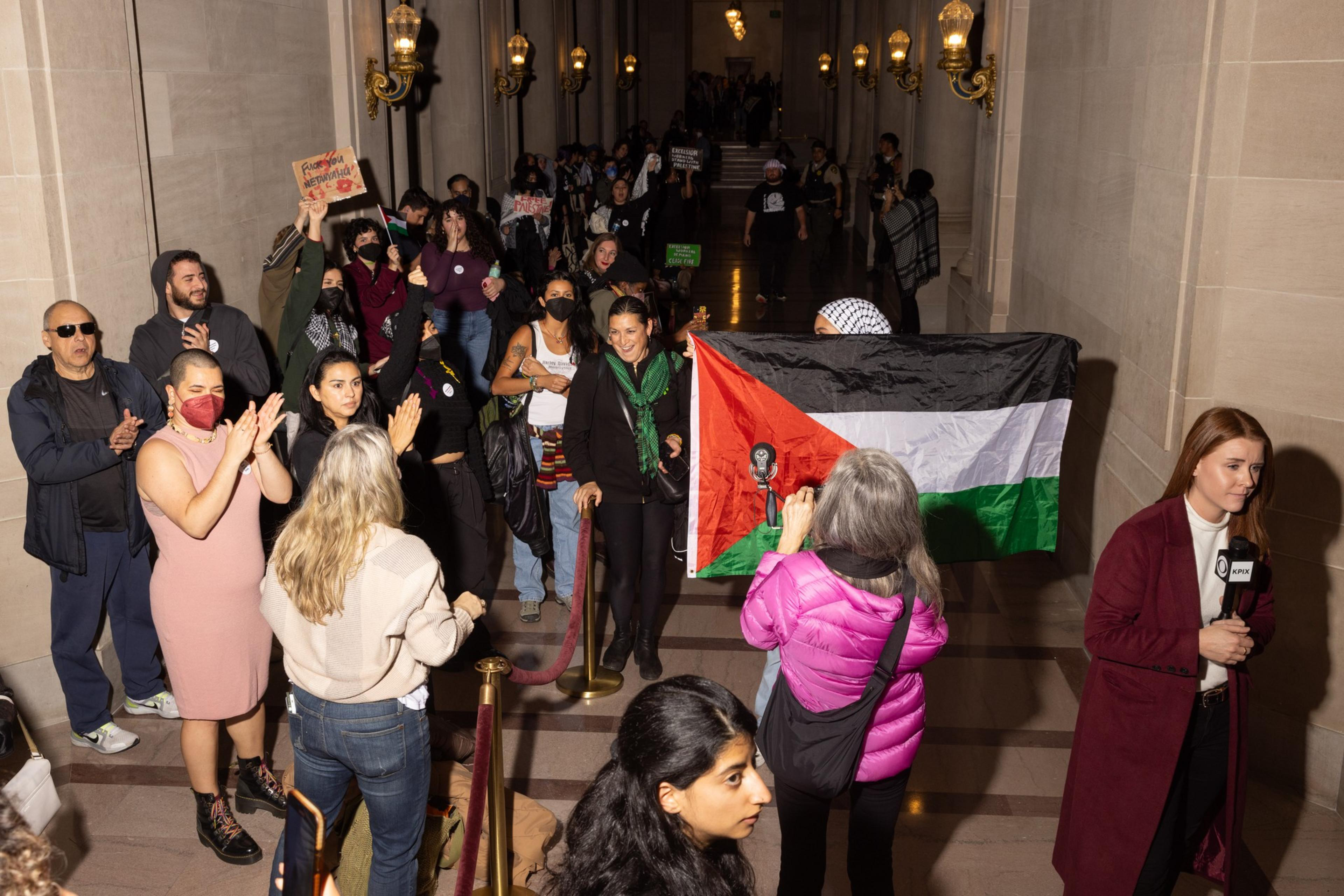 A long line out of people waiting to get in to the Board of Supervisors meeting with someone holding up a Palestine flag. 