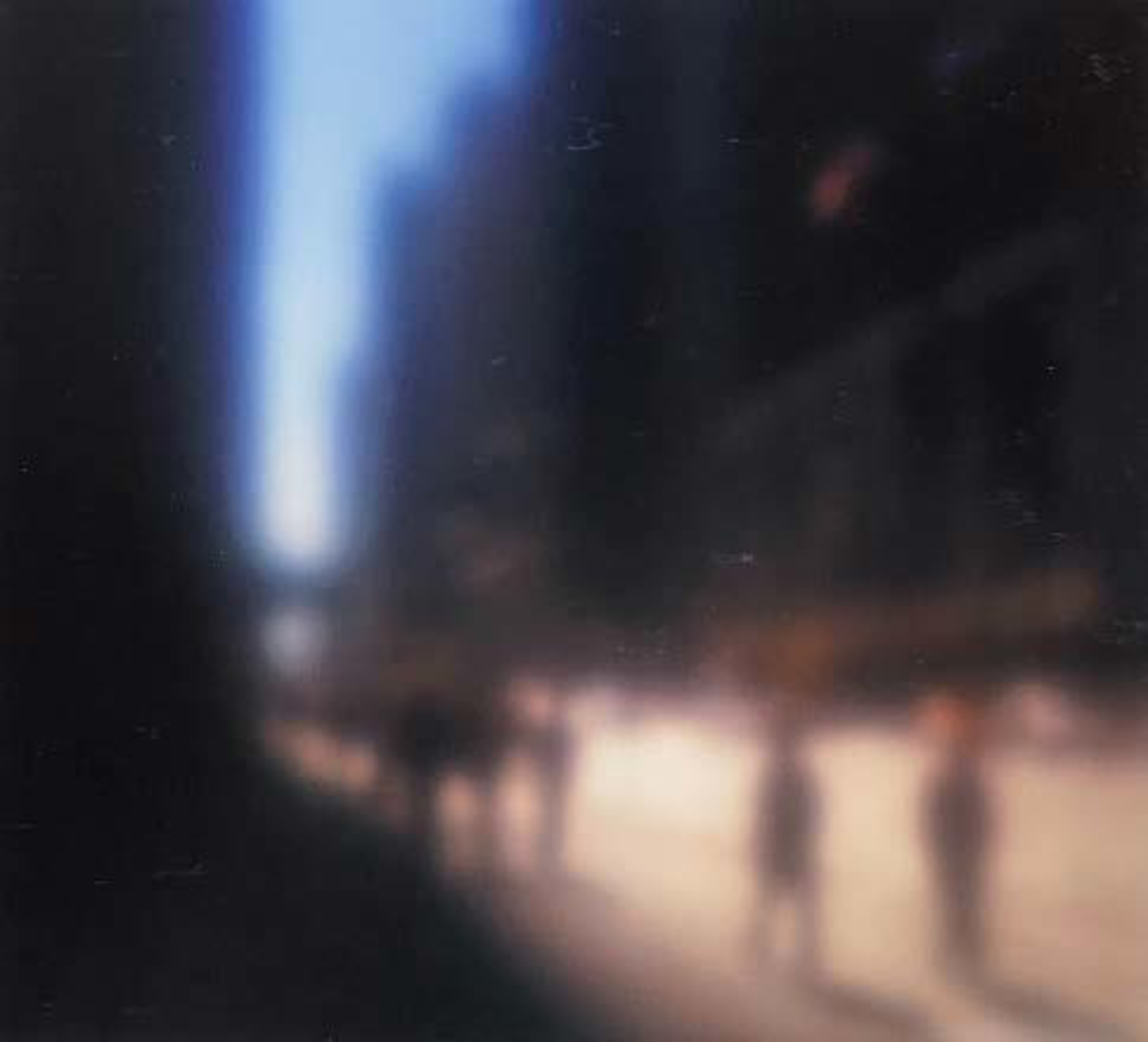 Silhouettes of people stand along a brightly lit street contrasted with a dark, shaded area under a tall building or structure.