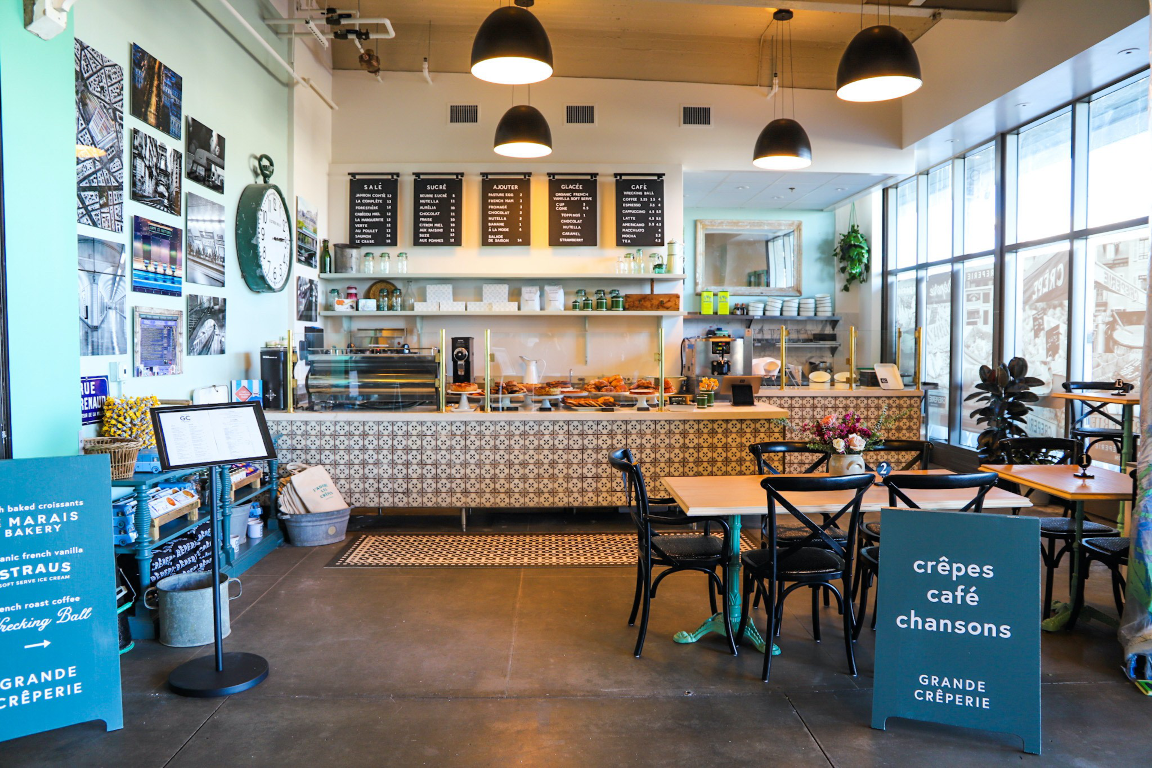 A bright café interior with tables, chairs, a patterned counter displaying pastries, menu boards, large windows, and a sign advertising crêpes, café, and songs.