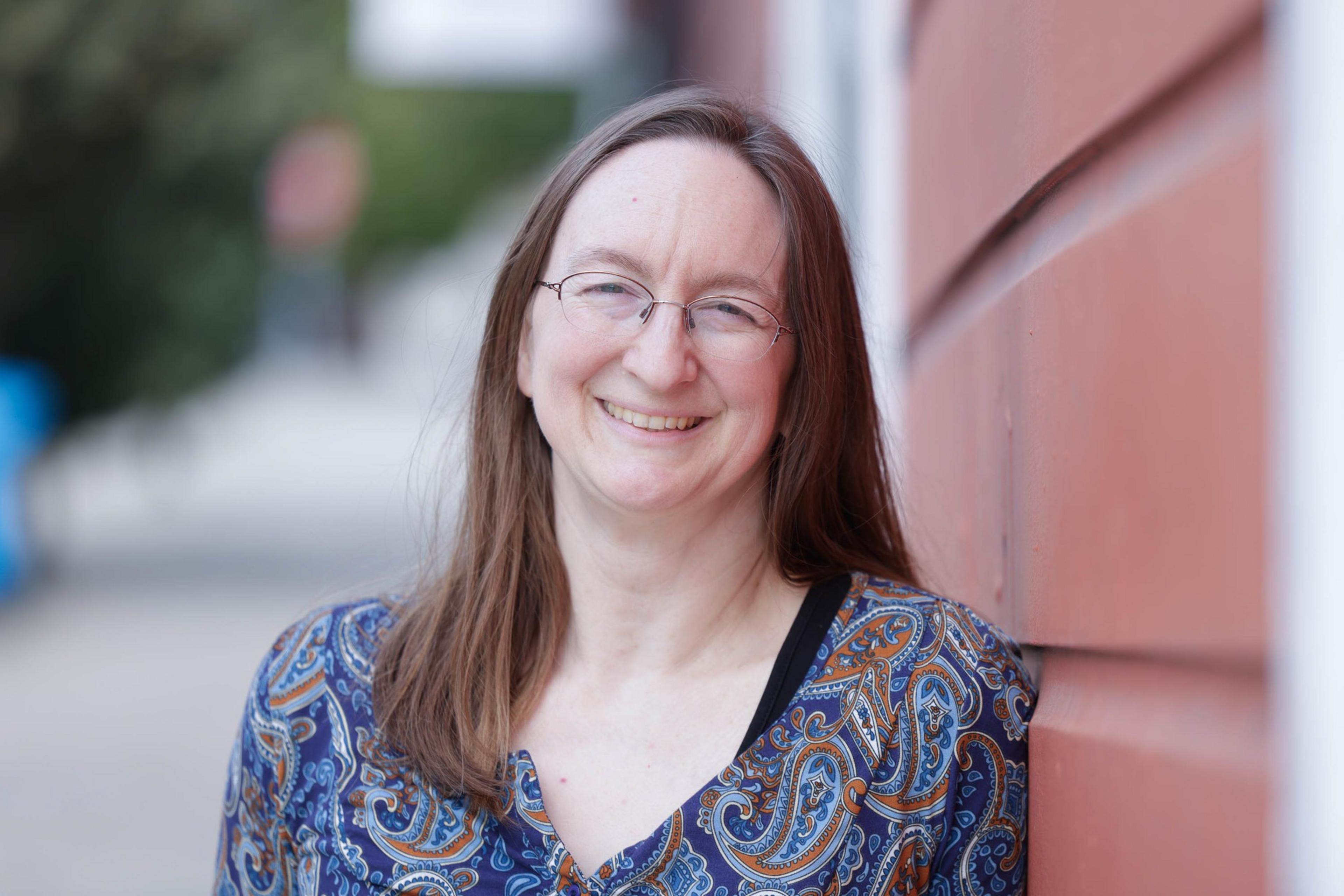 A smiling woman with glasses, wearing a patterned top, is leaning against a red brick wall.