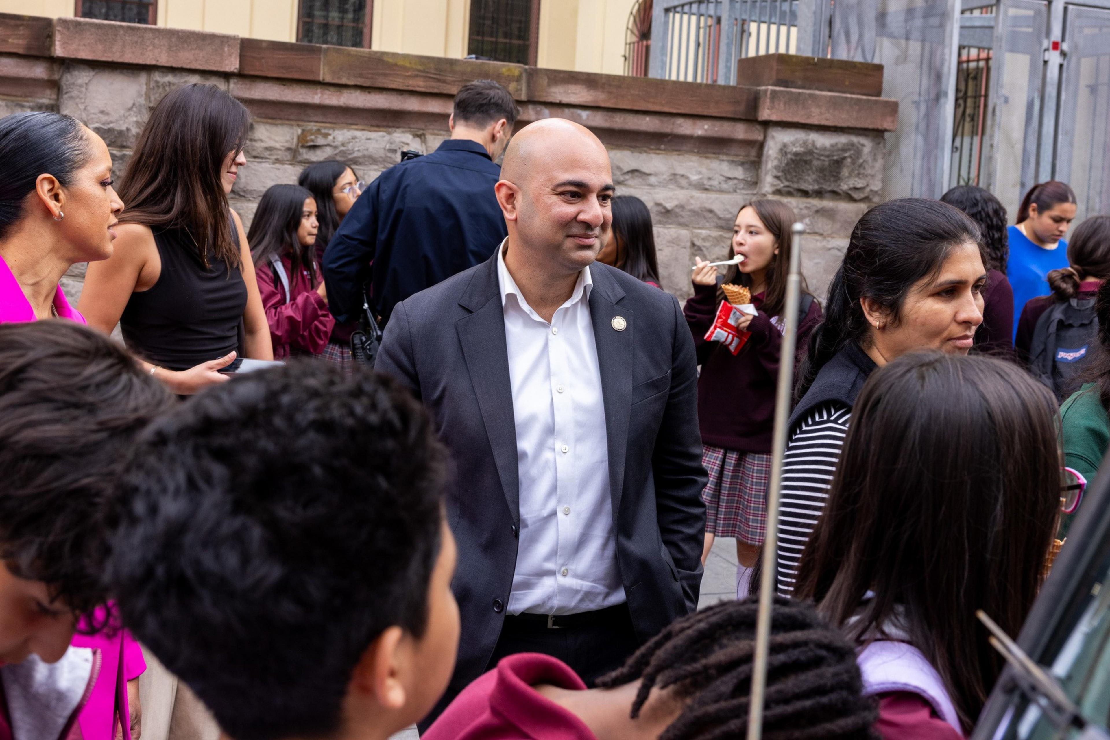 A bald man in a suit stands among a group of people, including children and adults, some of whom are eating or engaged in conversation outdoors.