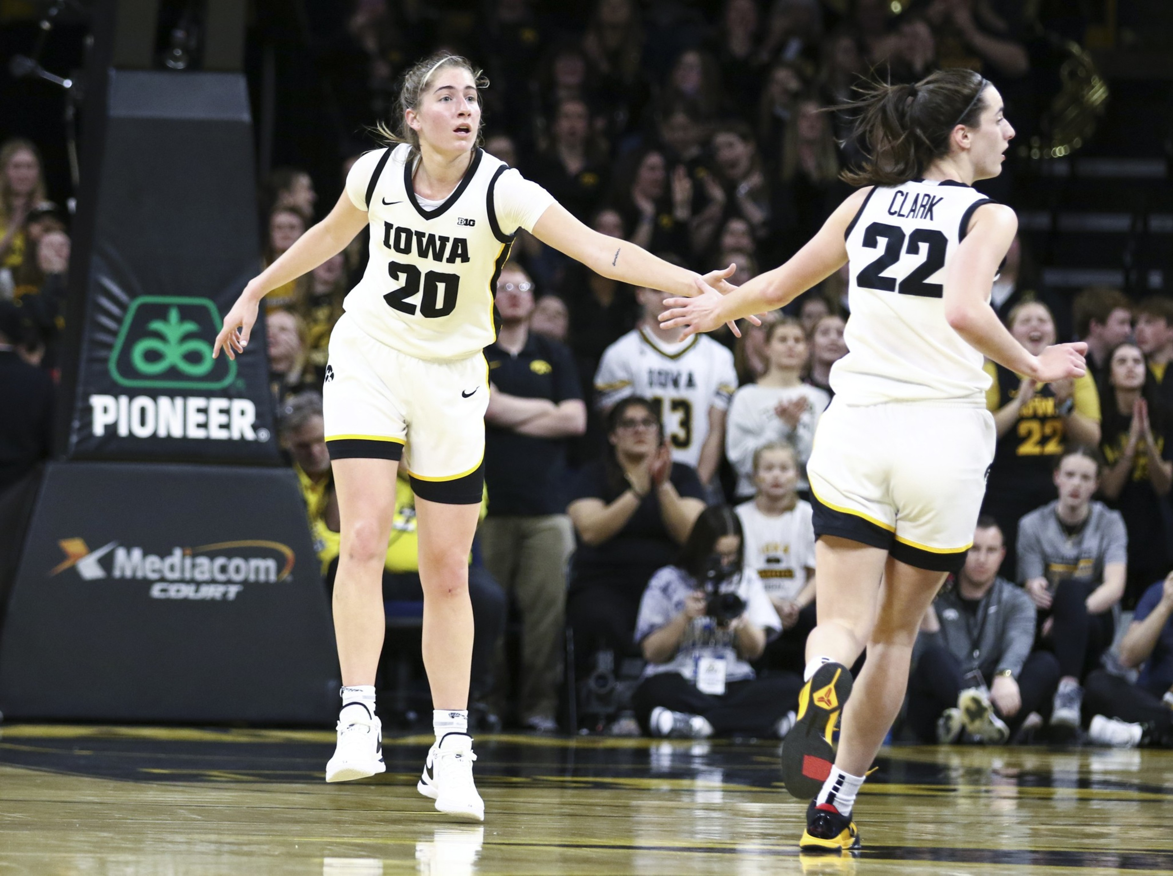 Two Iowa women's basketball players wearing white uniforms give each other a hand gesture during a game with a crowd in the background.