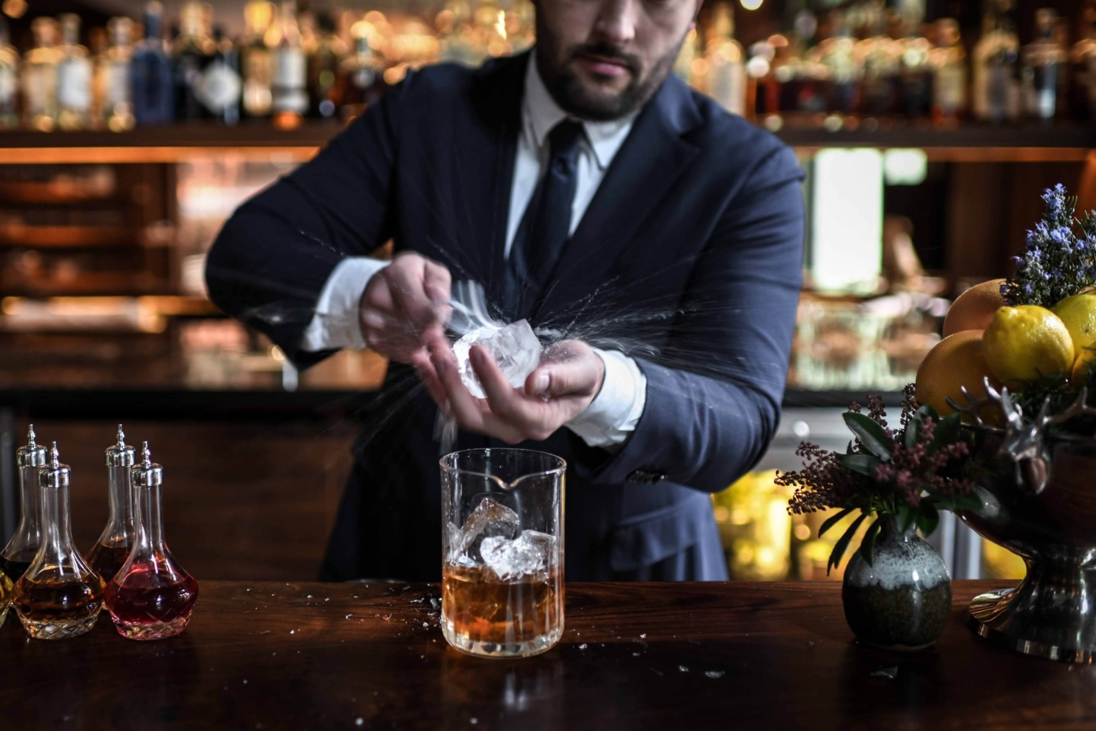 A bartender in a suit carves a large ice cube, creating splashes. Glass decanters with colored liquids and a citrus-filled vase adorn the bar counter.
