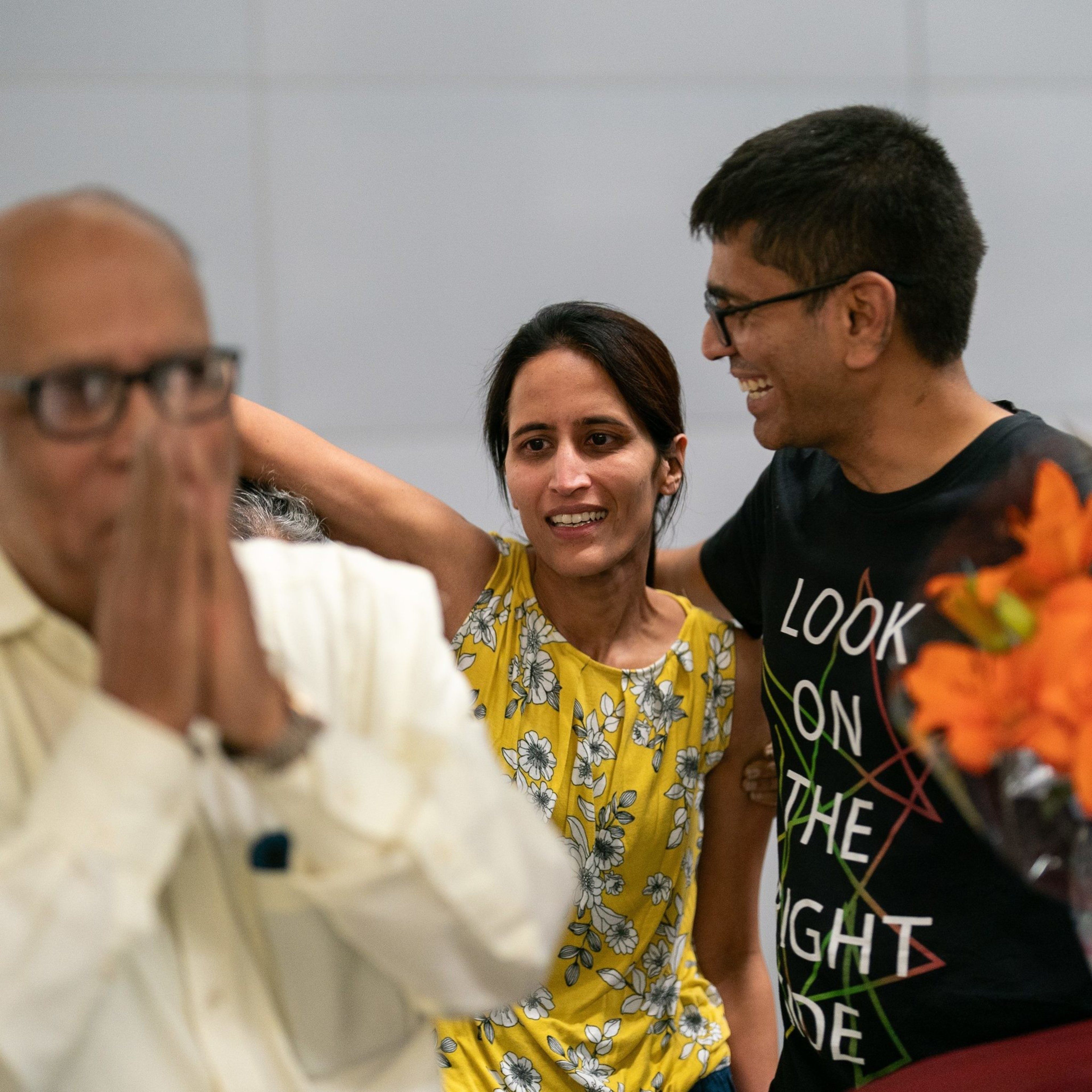 An elderly man in a white shirt stands with hands clasped in front of his face, while a woman in a yellow floral top and a man holding flowers smile and embrace.