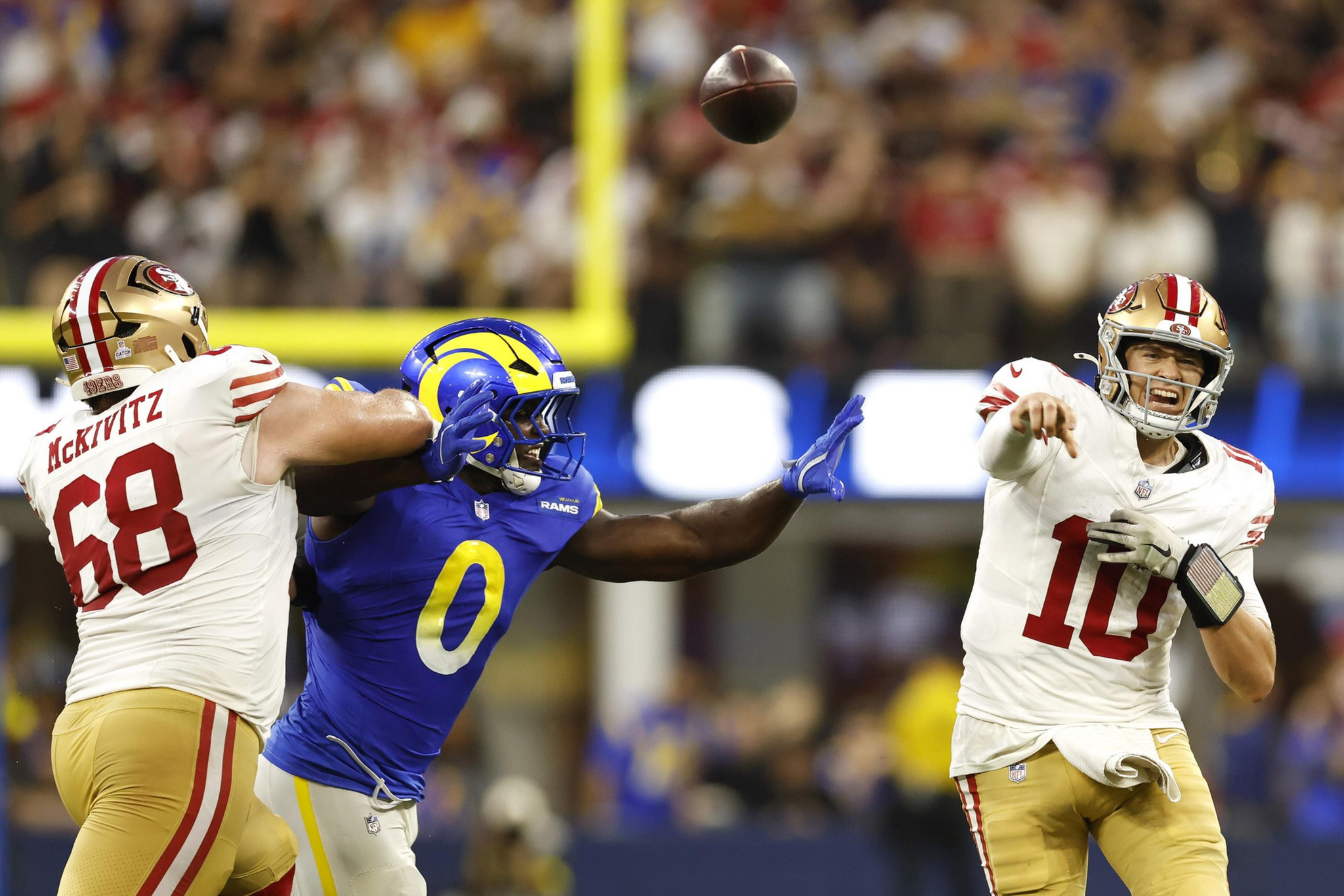 A football player in a white and red uniform throws the ball while a defender in blue and yellow tries to block, and another player in white blocks the defender.