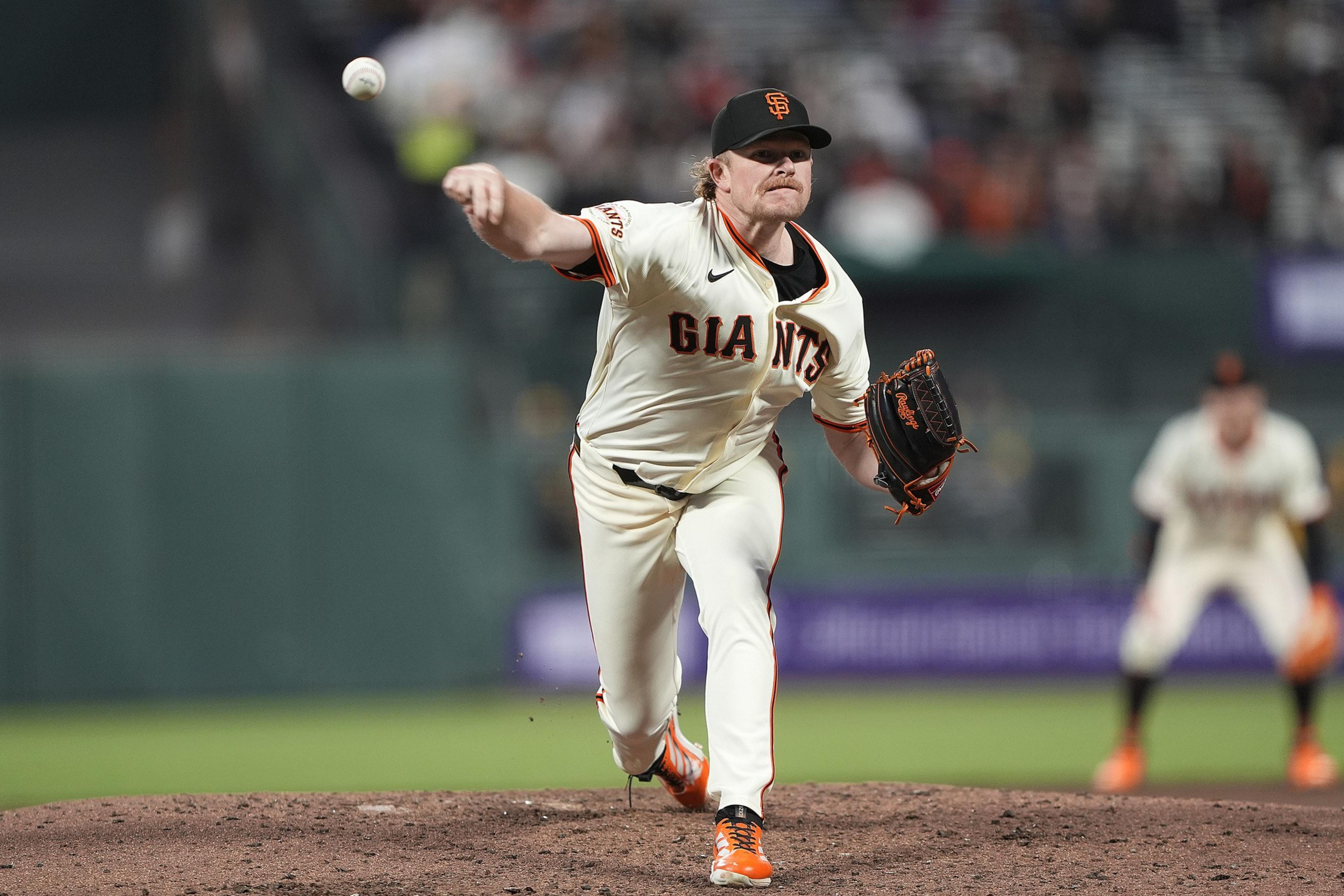 A baseball player in a white "Giants" uniform pitches a ball on the field. He's wearing an orange and black cap, with focused expression and motion.