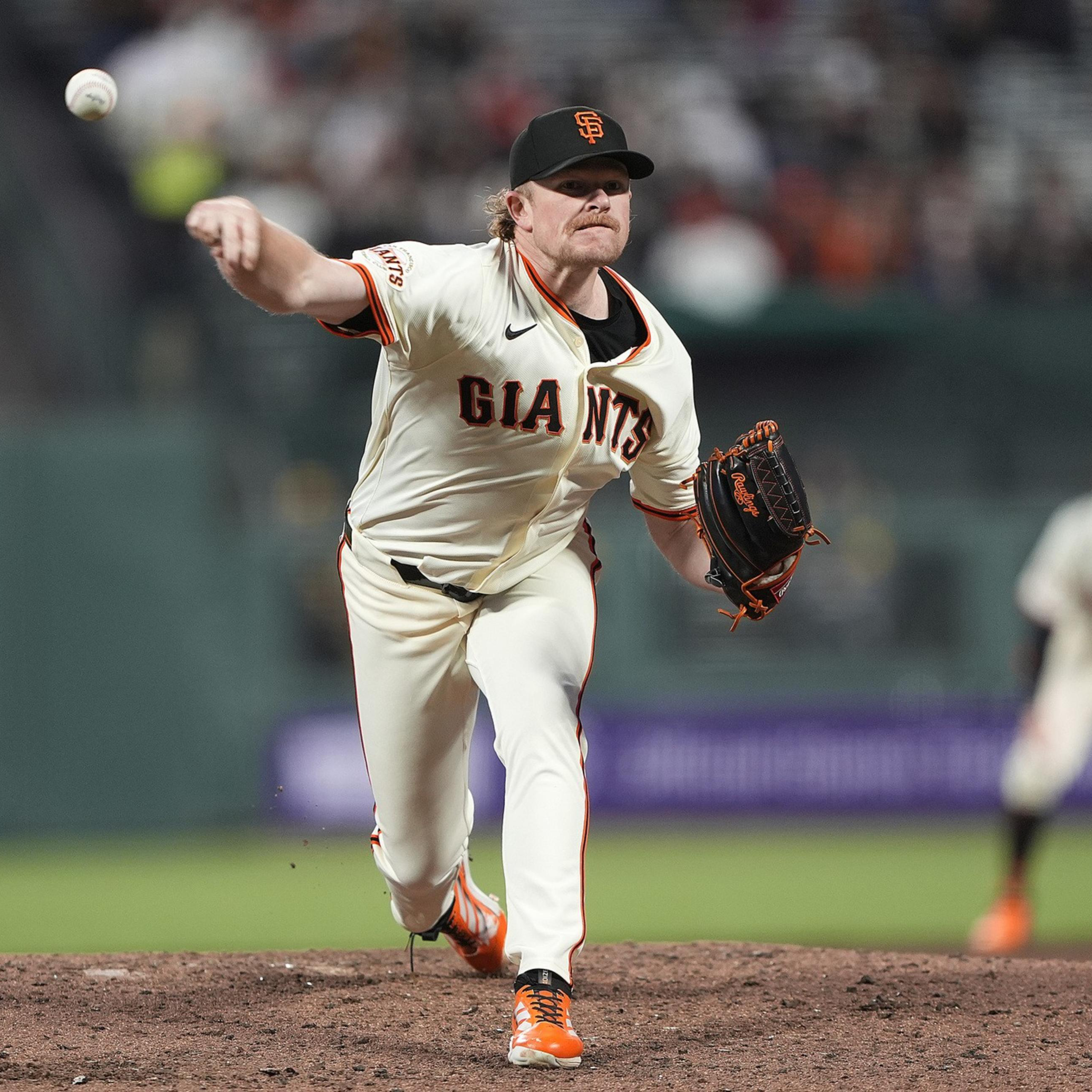 A baseball player in a white "Giants" uniform pitches a ball on the field. He's wearing an orange and black cap, with focused expression and motion.