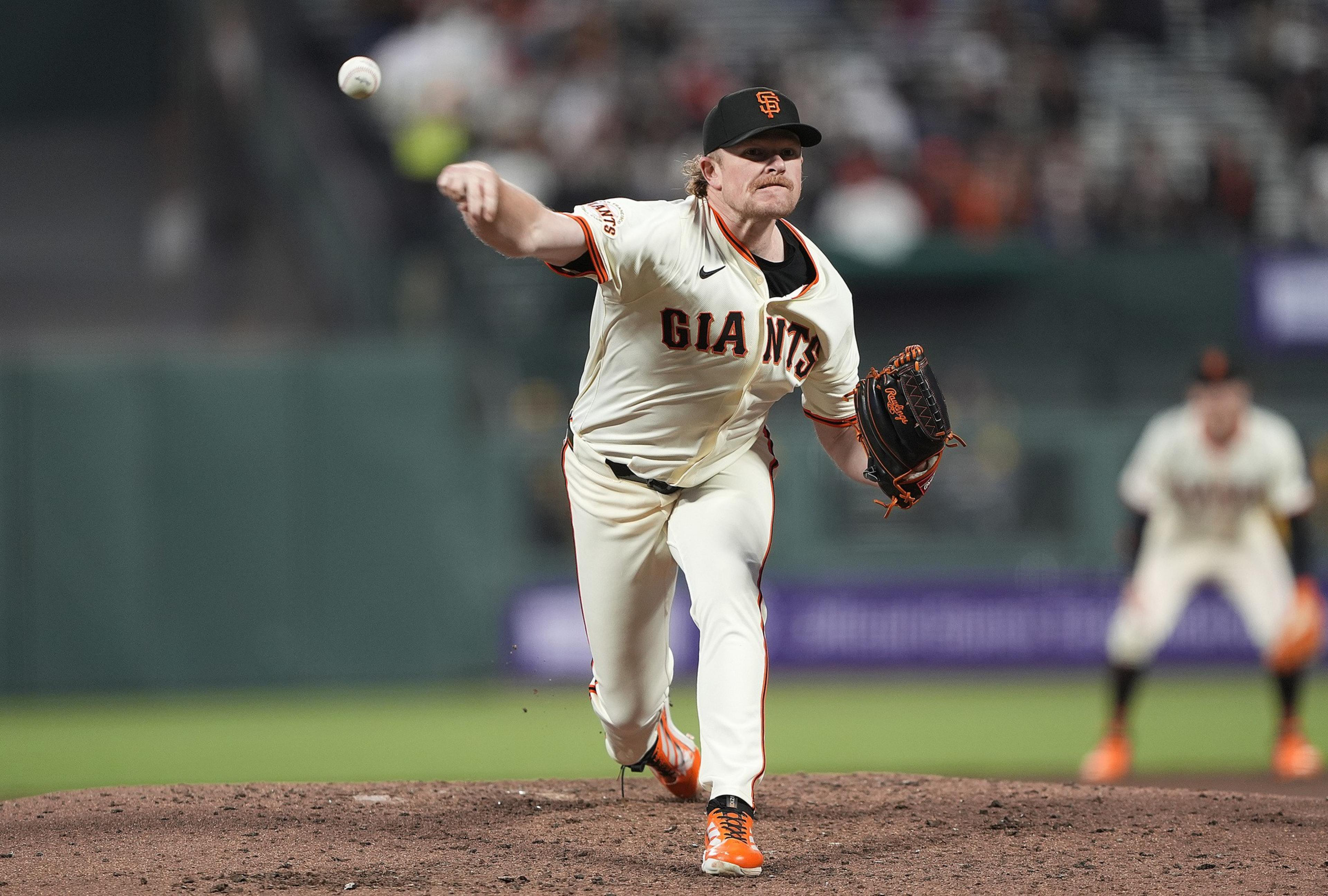 A baseball player in a white "Giants" uniform pitches a ball on the field. He's wearing an orange and black cap, with focused expression and motion.