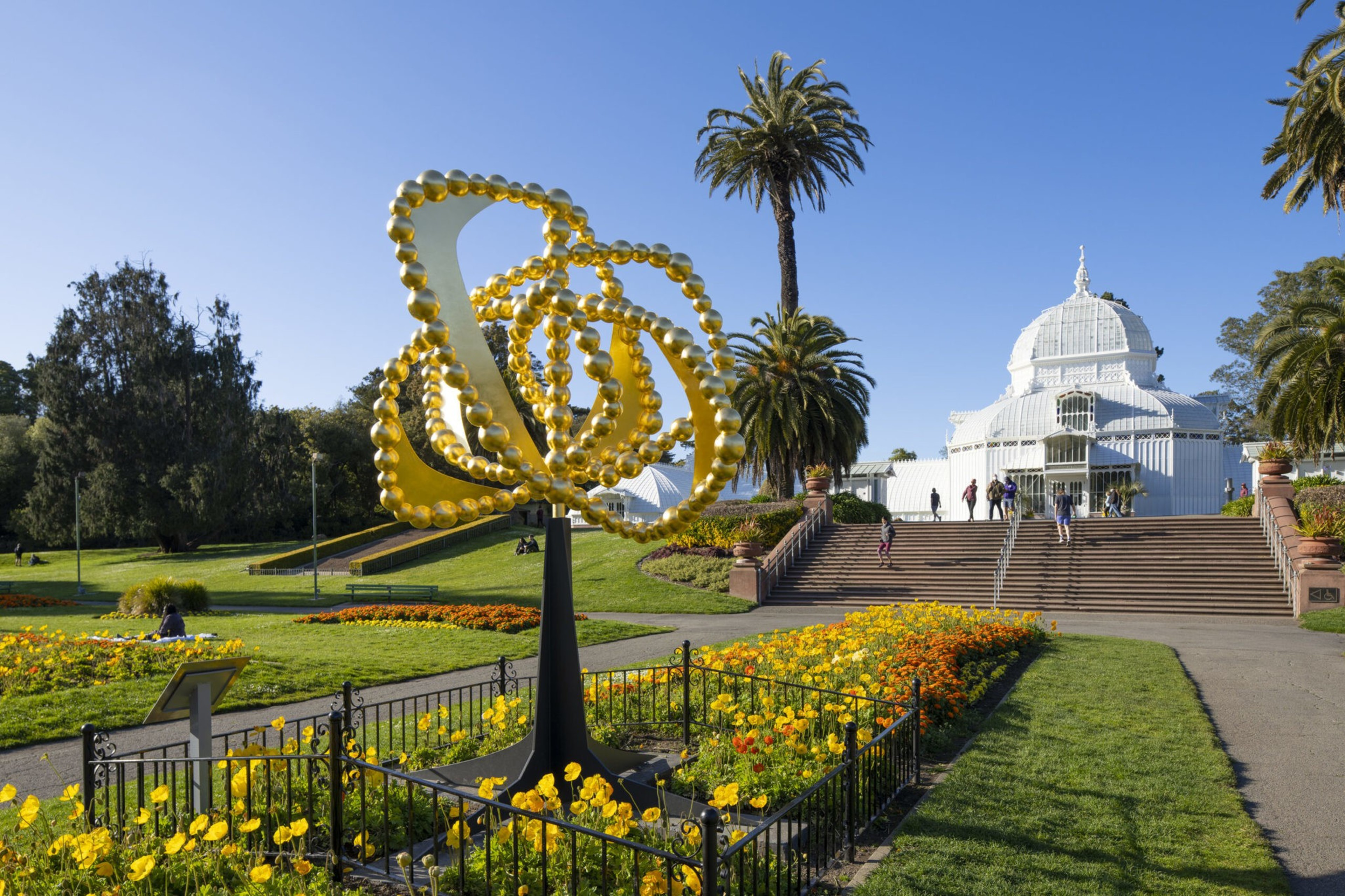 A bright yellow abstract flower sculpture stands amidst yellow flowers with palm trees and a white domed conservatory in the background.