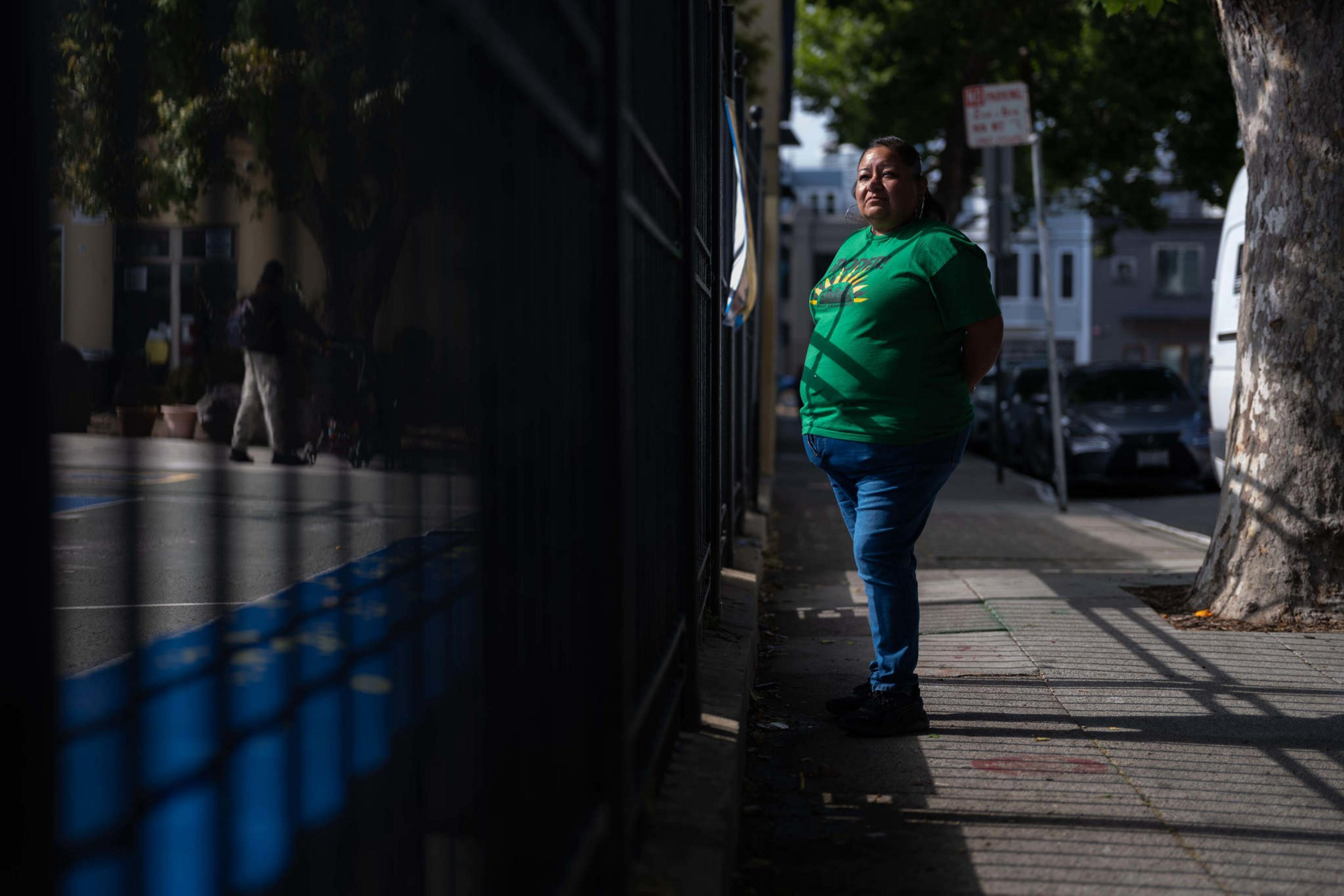 A person in a green shirt stands on a shaded sidewalk, looking ahead. A fence casts shadows, and another person is visible in the background.