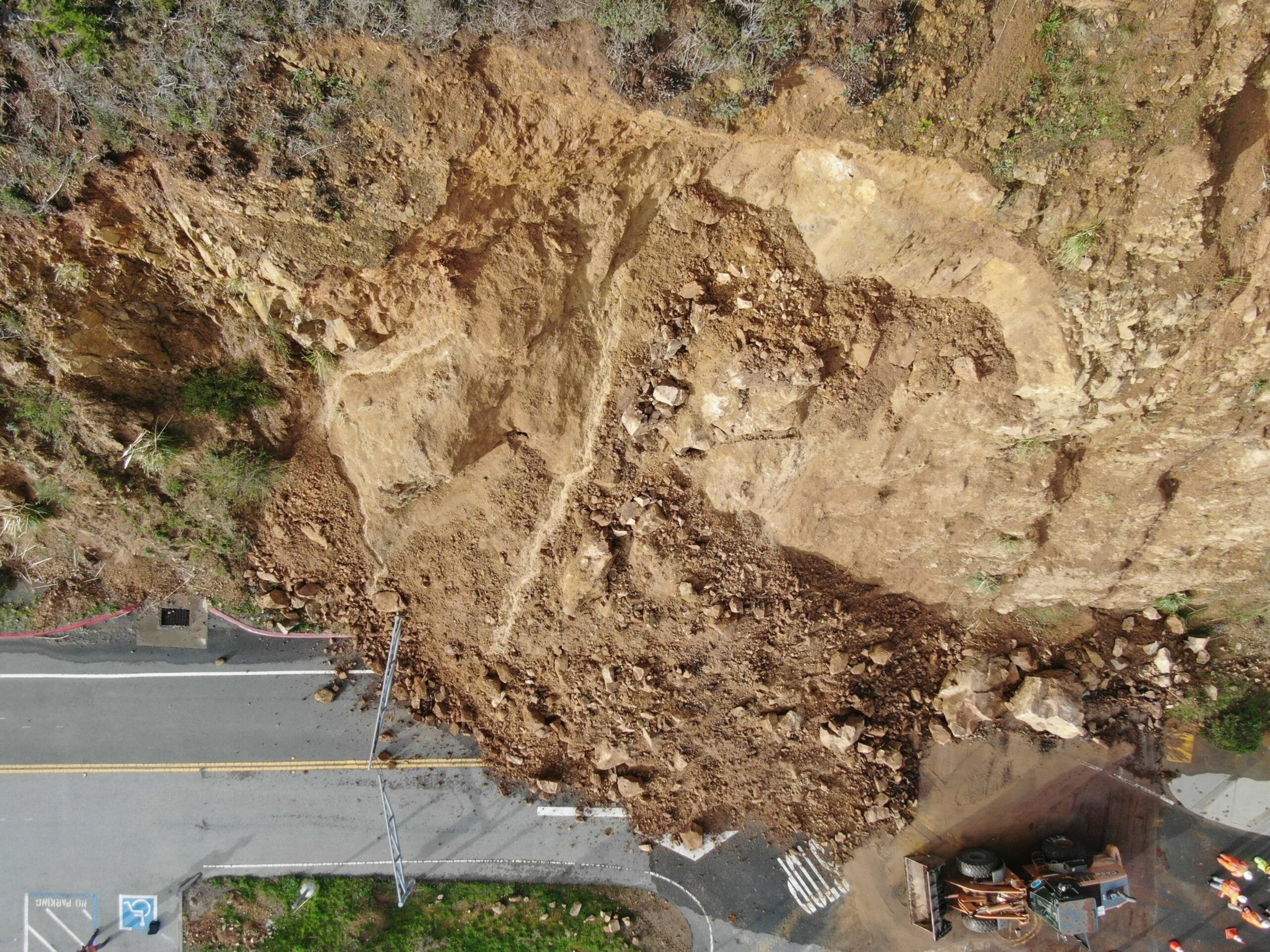 A large landslide has covered most of a two-lane road with dirt, rocks, and debris, blocking passage and causing a construction vehicle to stop.